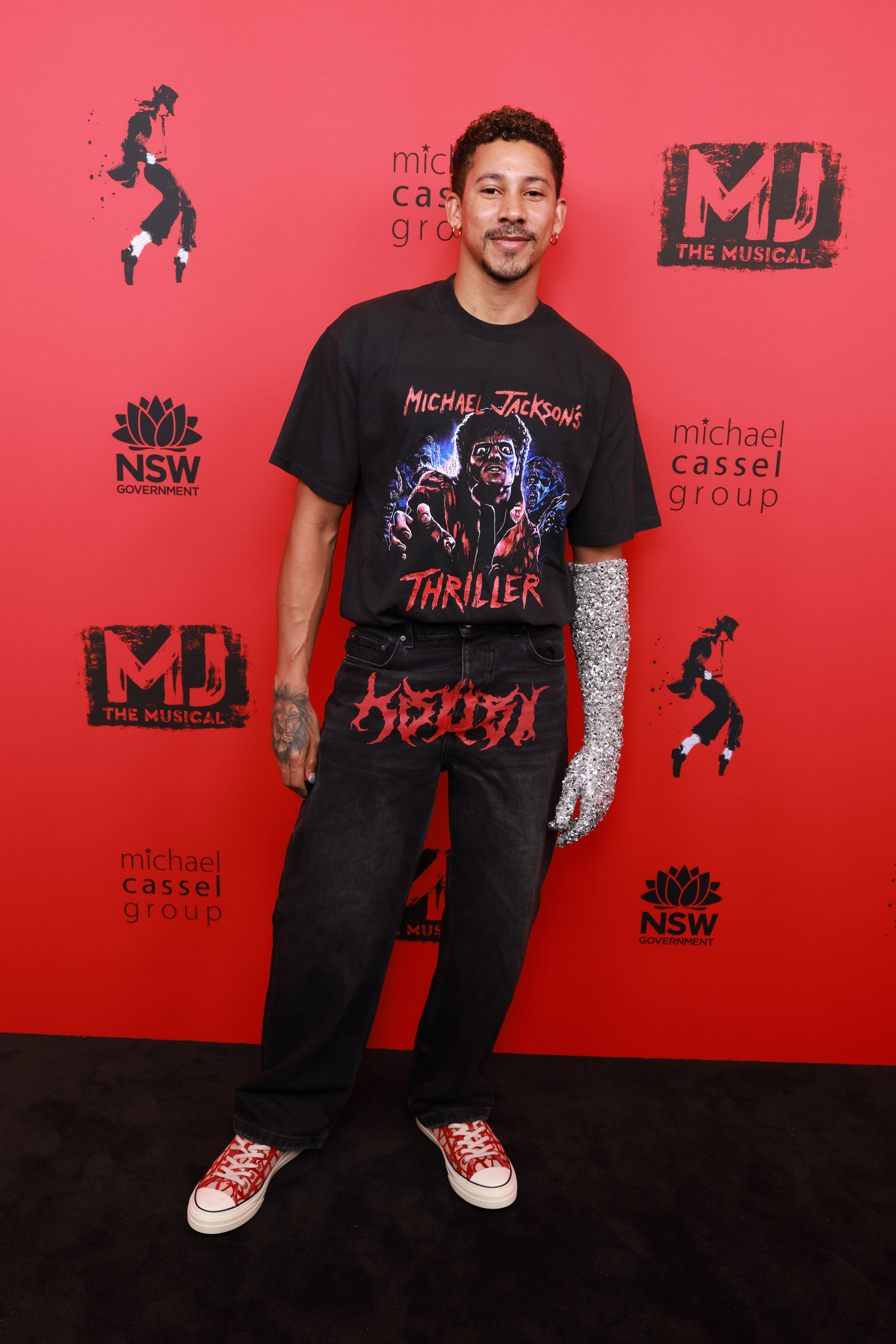 Person wearing a graphic t-shirt and jeans with a shimmering glove on one hand, posing on a red carpet at an event for "MJ The Musical."