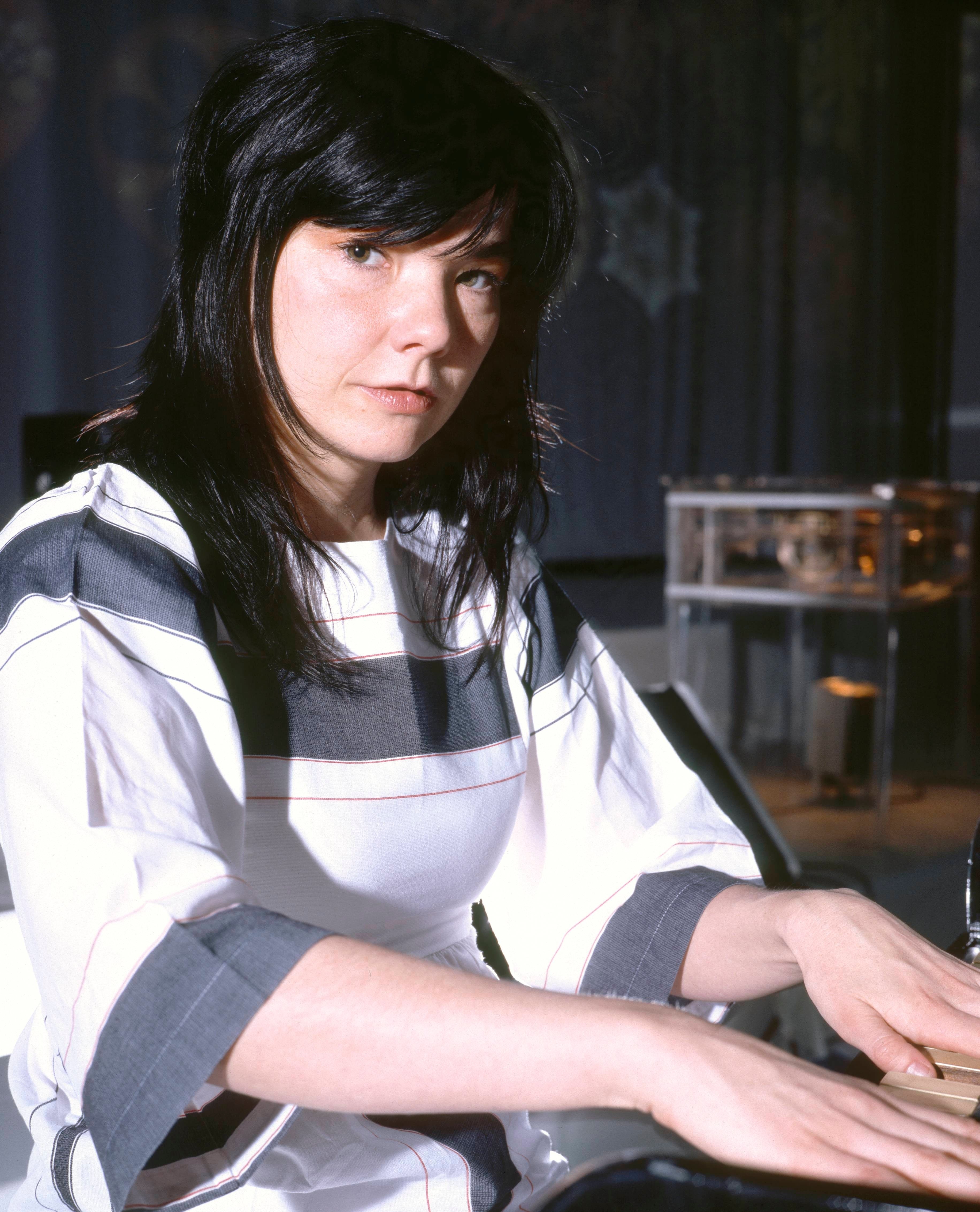 Person playing the piano, wearing a patterned dress, looking at the camera. The setting is indoors with a modern backdrop