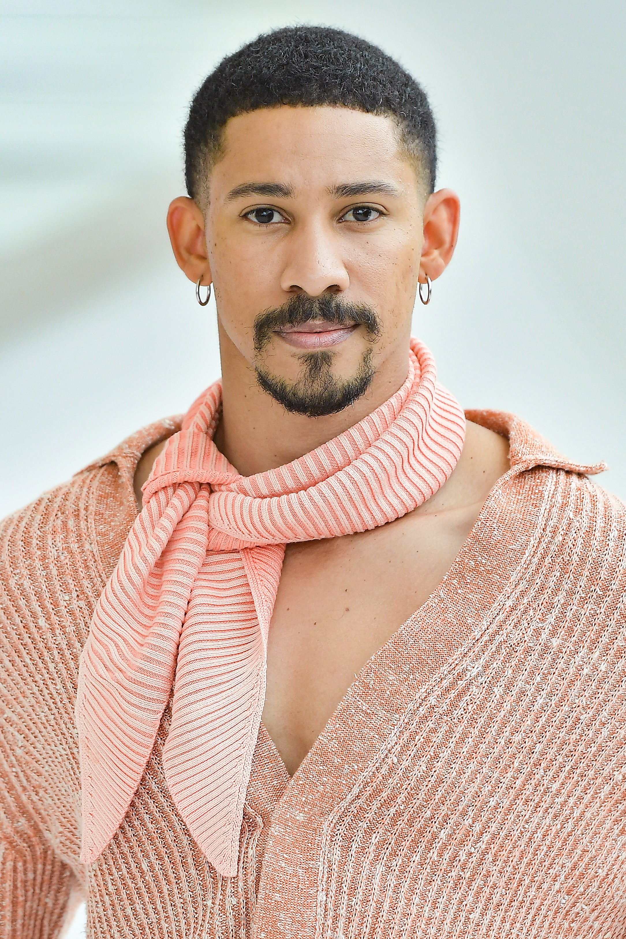 Person with short hair, hoop earrings, and a textured open shirt, styled with a scarf around the neck, poses confidently