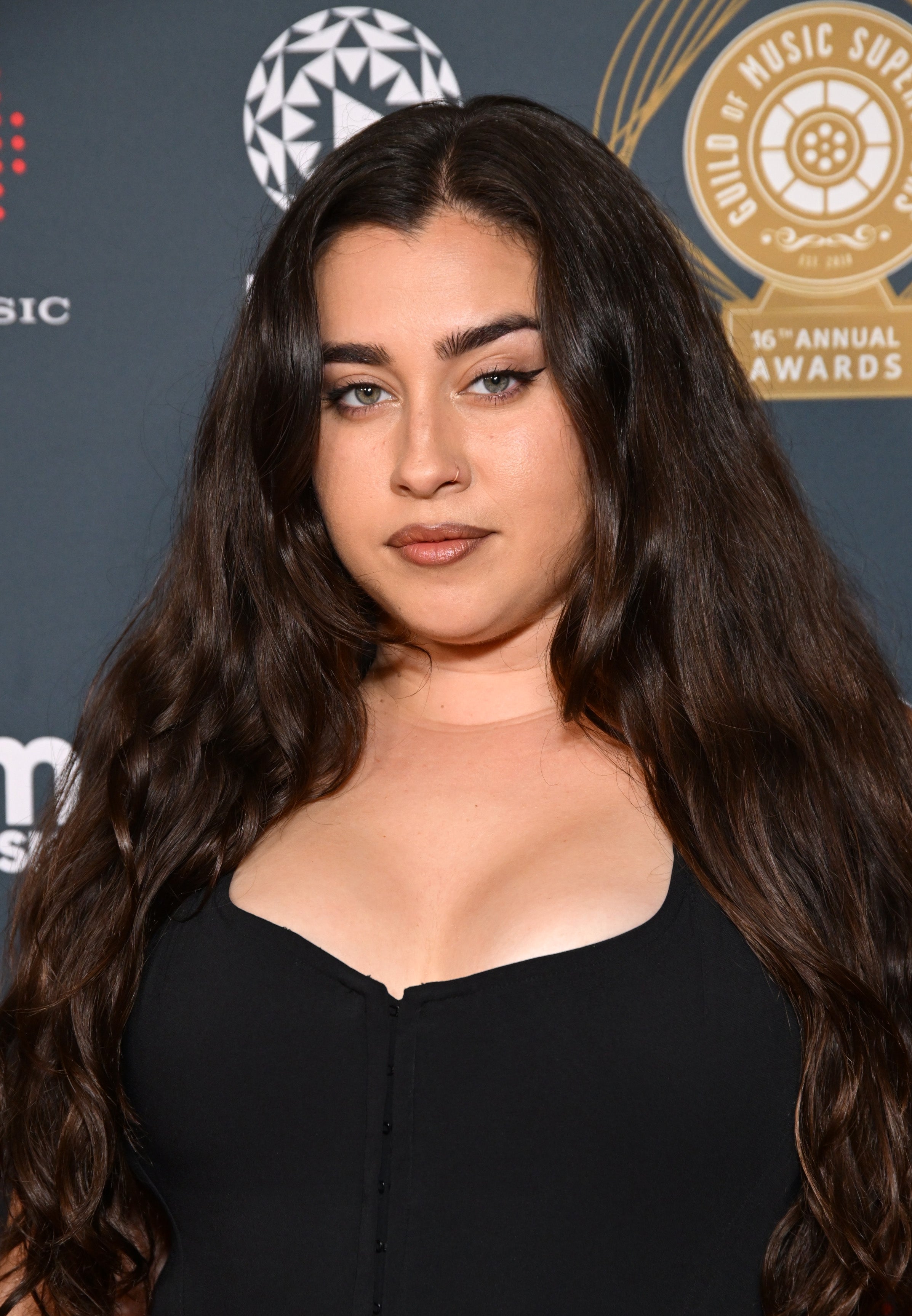 Person with long hair in a stylish, fitted black outfit poses at a music event. Background includes award logos and text