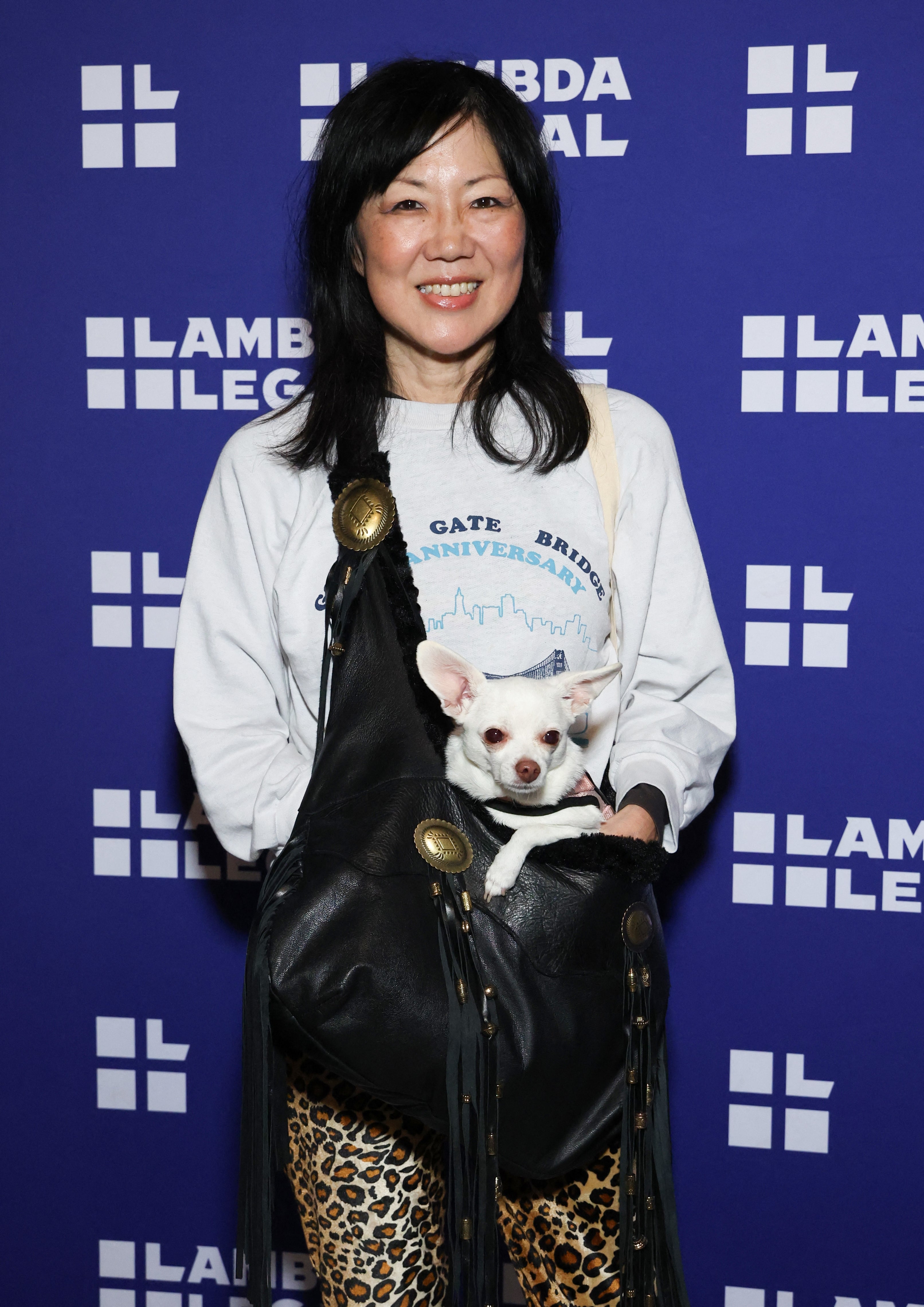Person smiling at an event, wearing a graphic sweatshirt and leopard print pants, holding a small dog in a bag. Lambda Legal backdrop