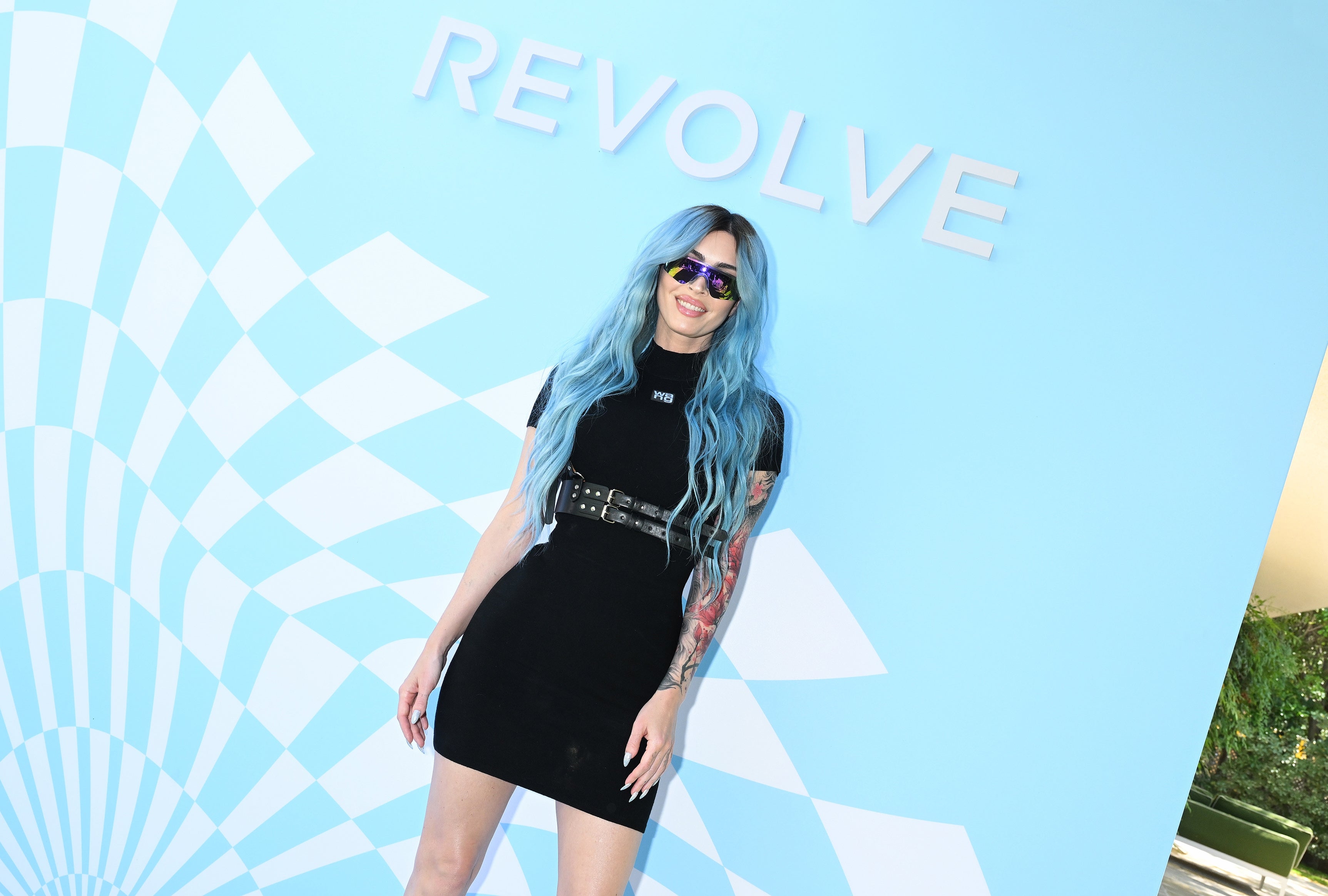 Person with long blue hair in a stylish black dress and sunglasses, standing in front of a Revolve backdrop
