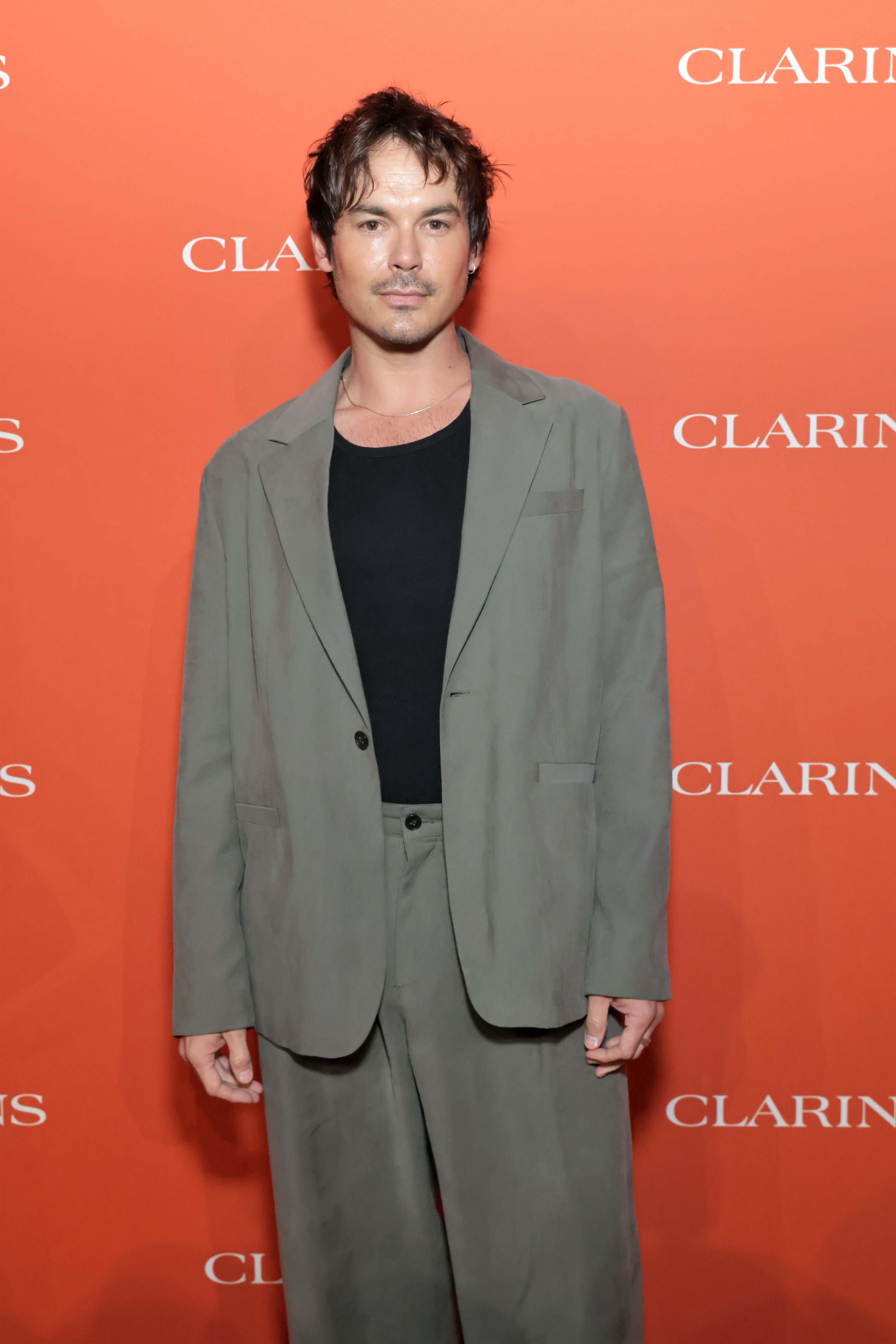 Person wearing a loose-fitting gray suit and black shirt, standing on a red carpet with a branded backdrop at a Clarins event
