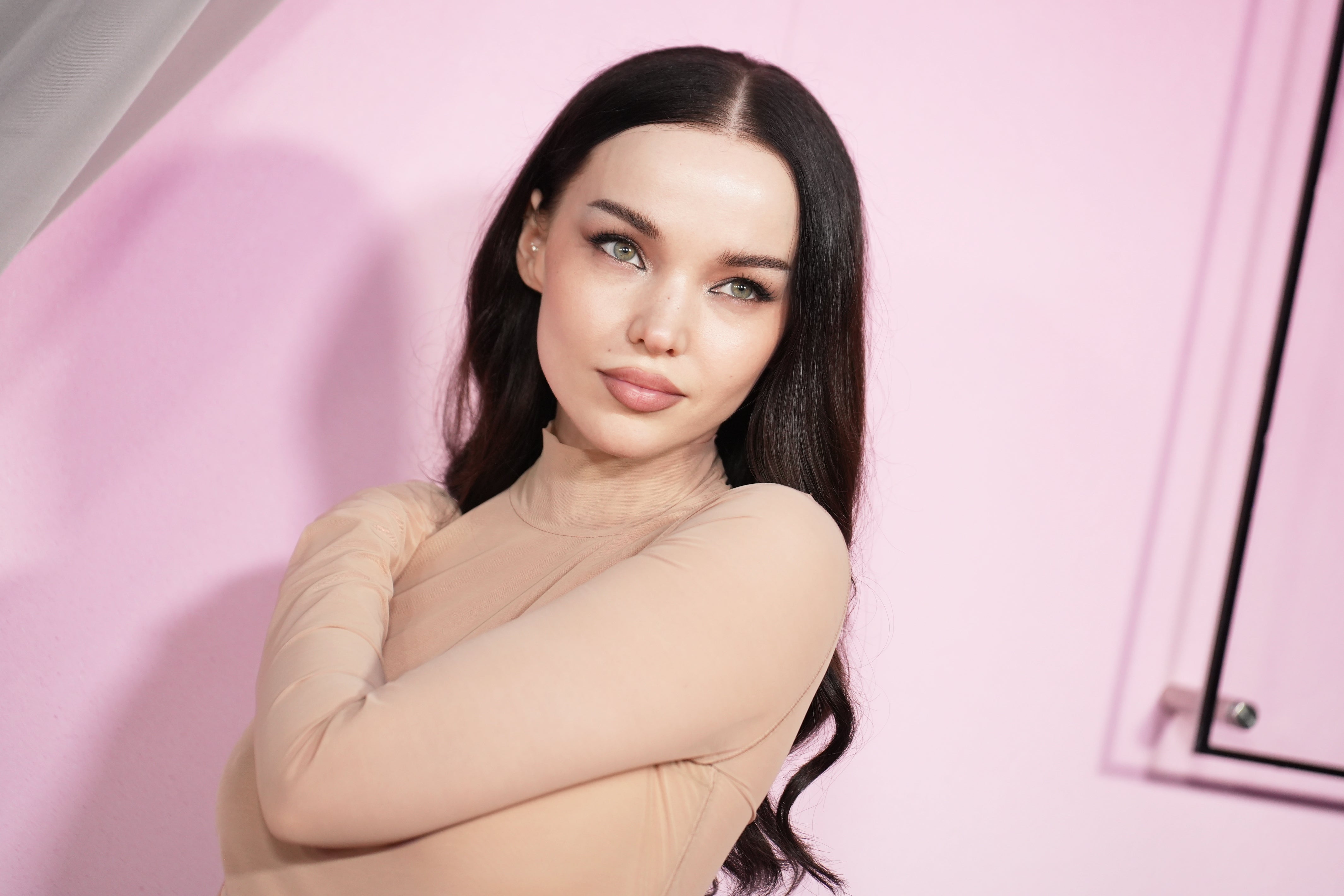 Person with long, dark hair stands against a pink background, wearing a form-fitting outfit, looking confidently at the camera