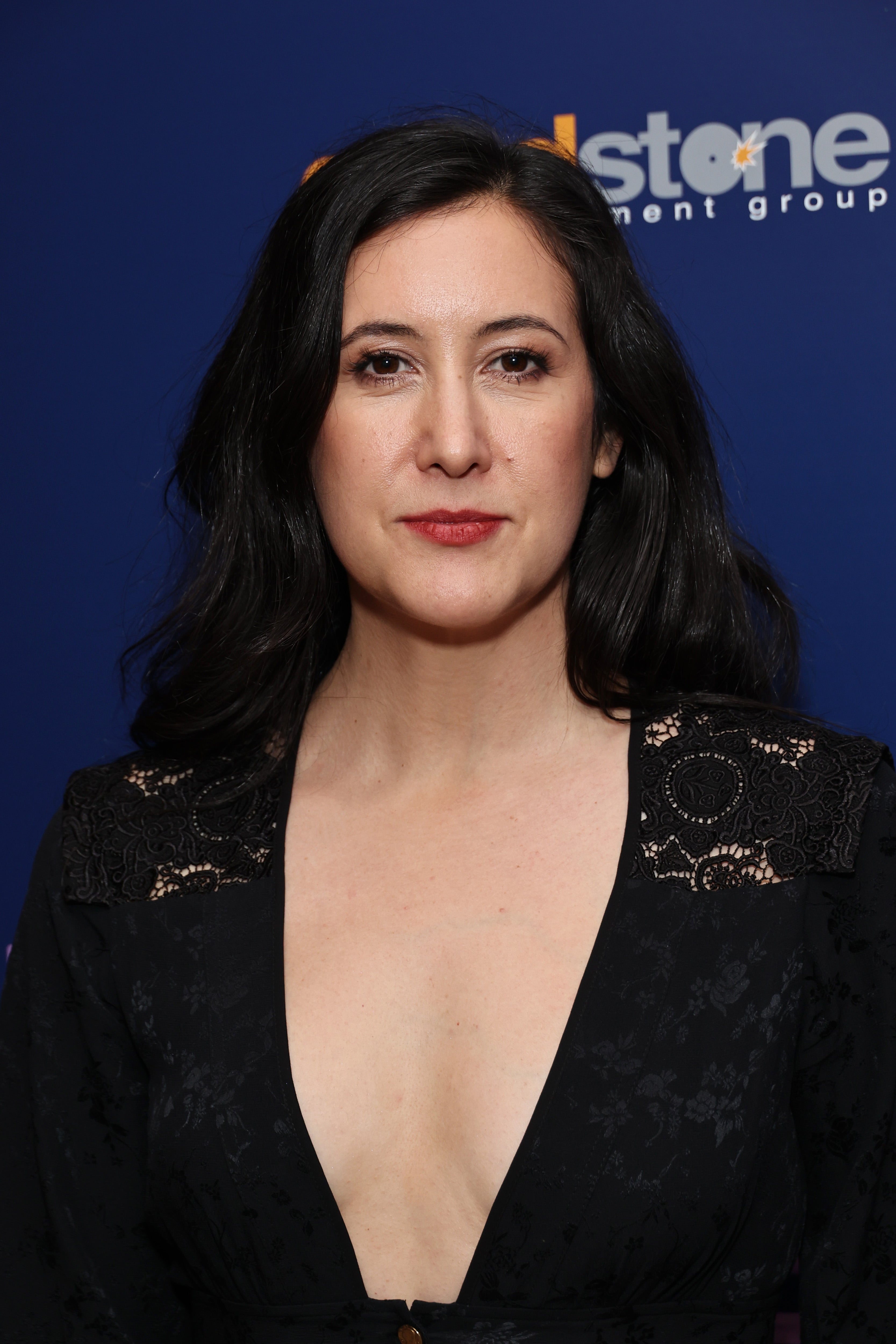 A person in a deep V-neck black dress poses on the red carpet, their expression serene and composed