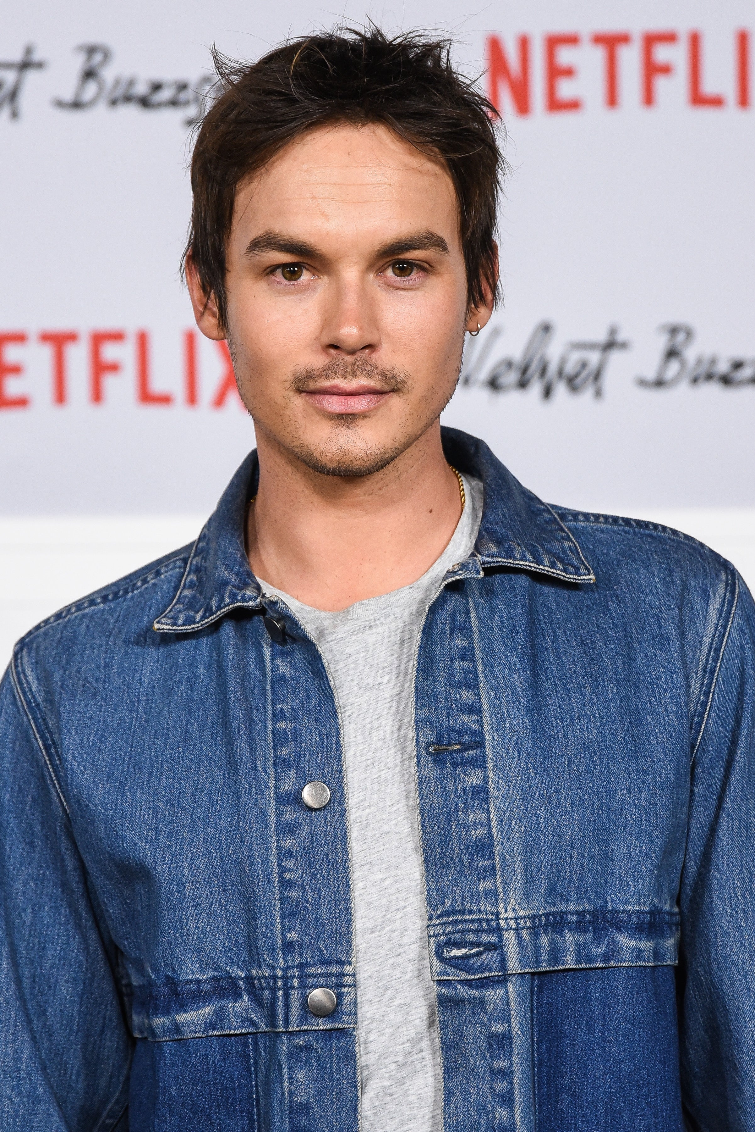 Person in a denim jacket and gray shirt at a Netflix event backdrop. Short hair, slight facial hair