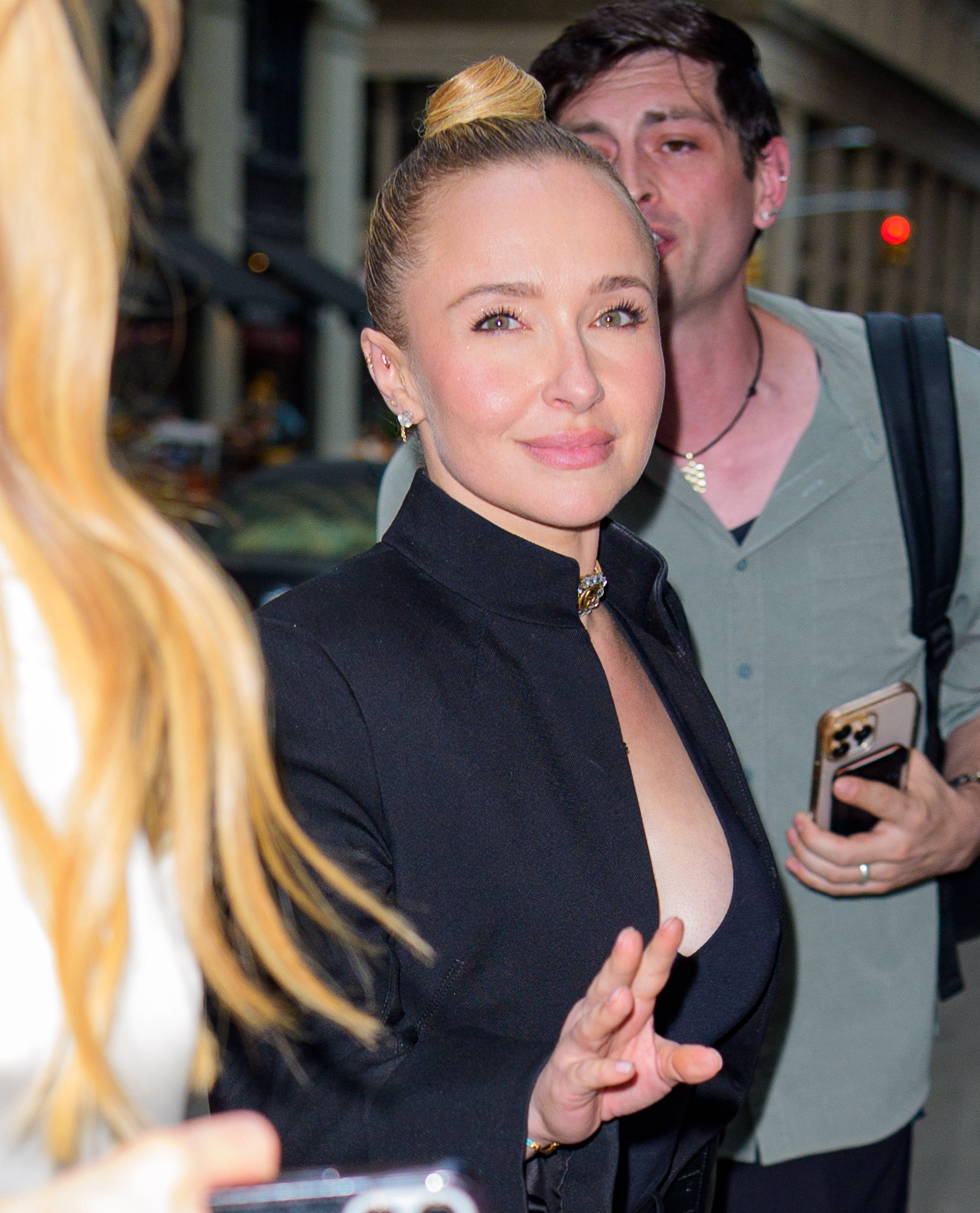 Person in a sleek black outfit with a plunging neckline poses confidently, holding up a hand with a warm expression. Another person stands nearby