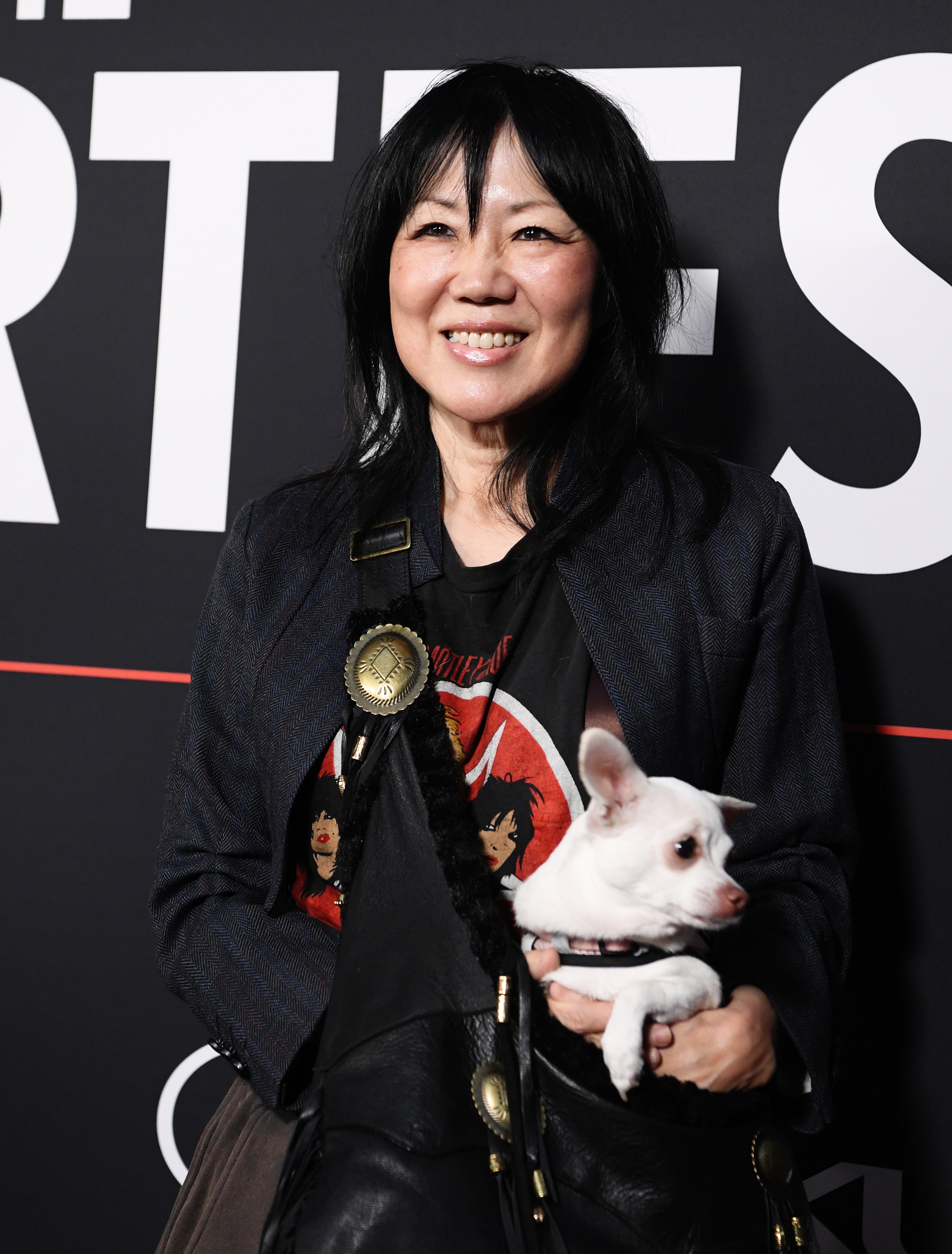 A person in a casual jacket and graphic t-shirt holding a small dog, posing at an event, with a confident smile