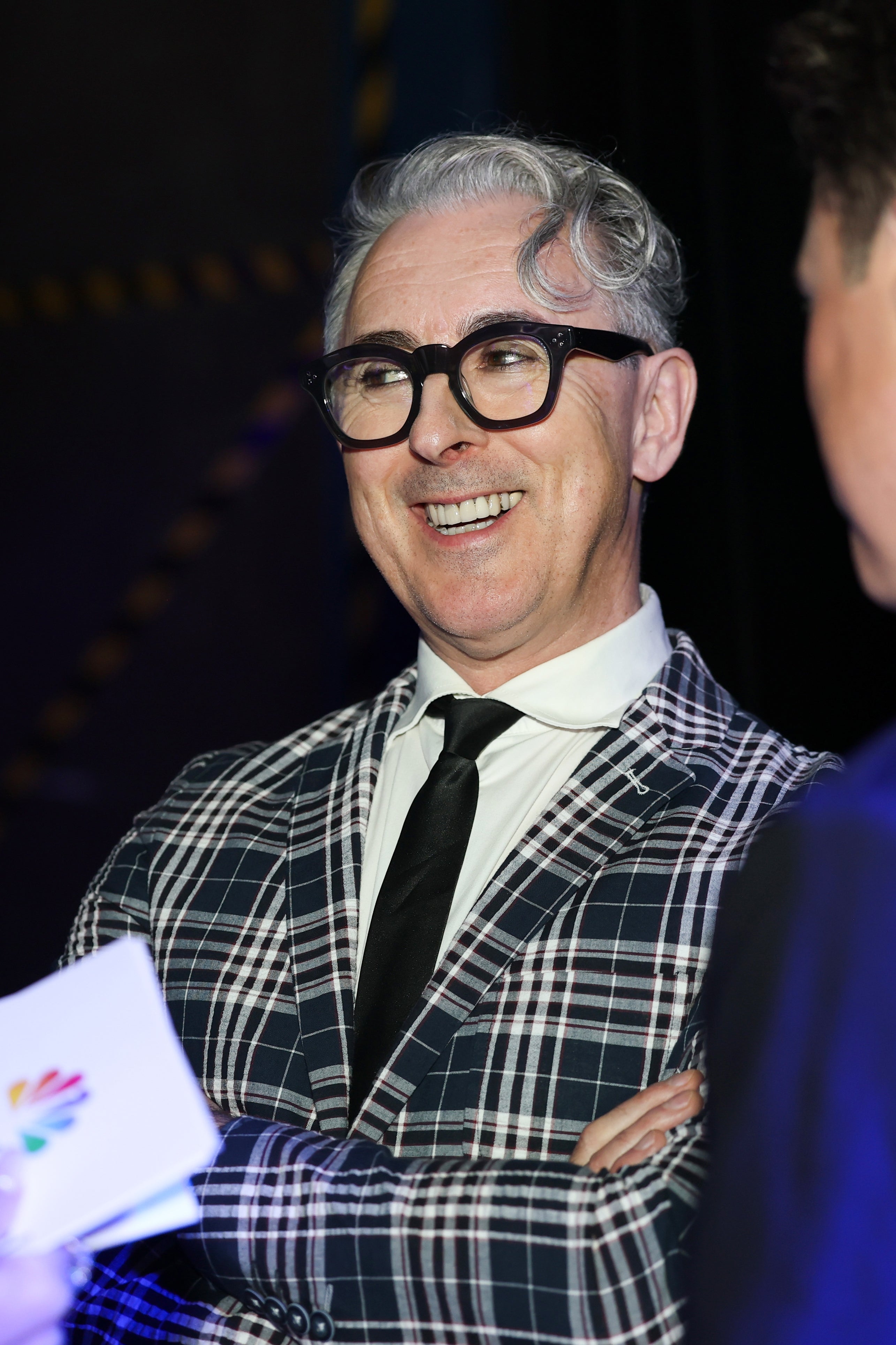 Person in a patterned suit and tie, smiling warmly at an event, engaged in conversation