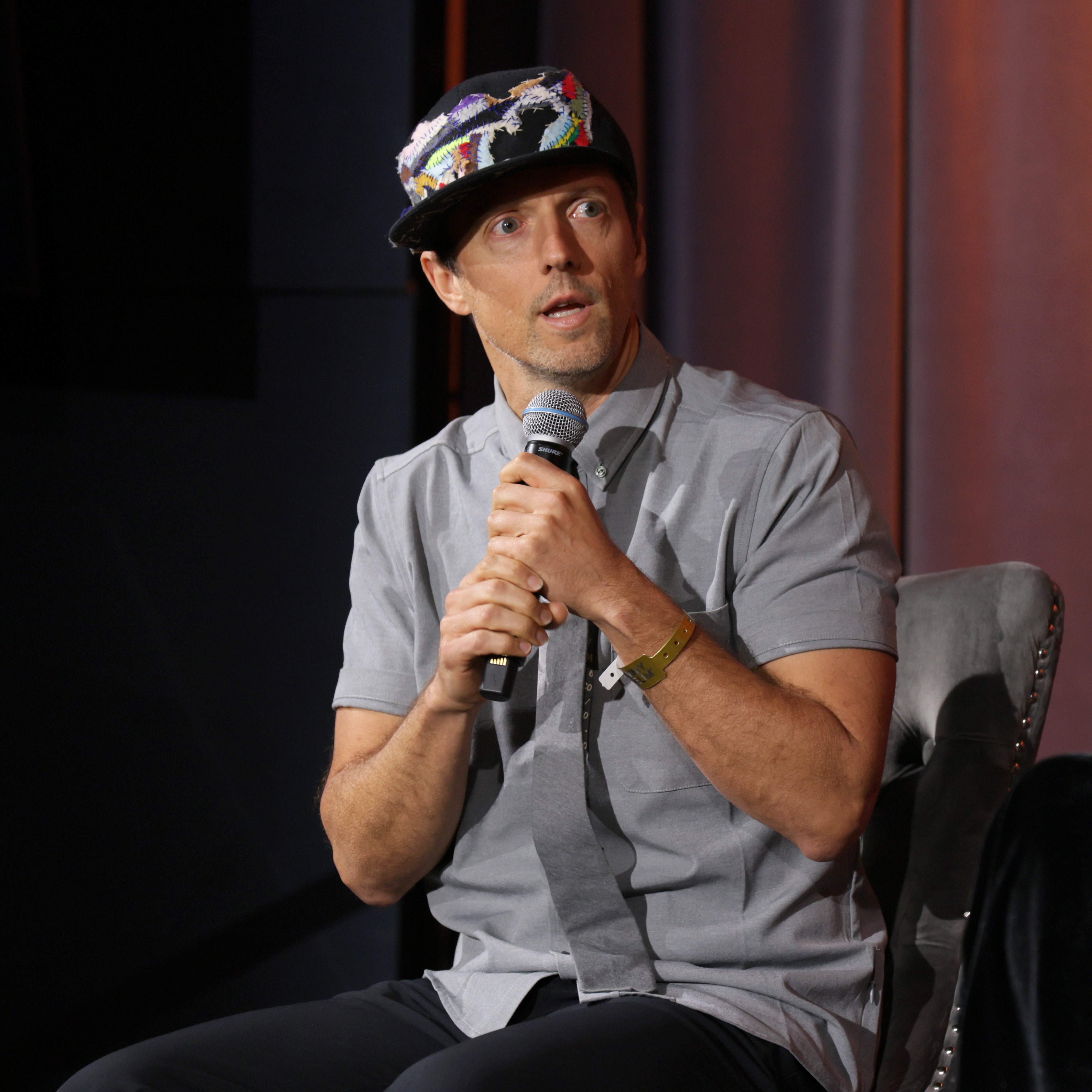 A person with a colorful cap and casual outfit speaks into a microphone while seated, appearing engaged in an event setting