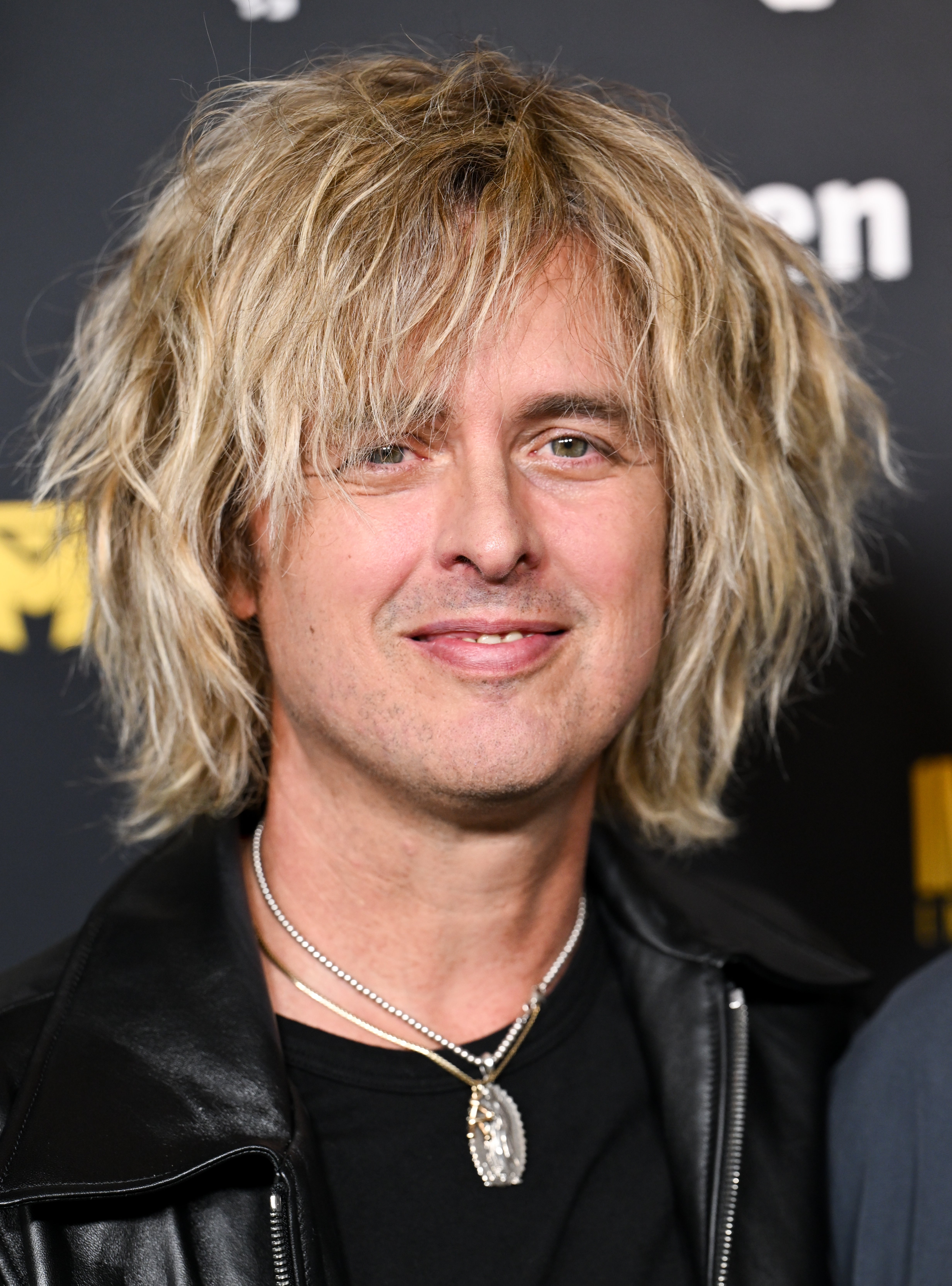 Person with tousled, shaggy hair in a black leather jacket, wearing a silver necklace, smiling at an event