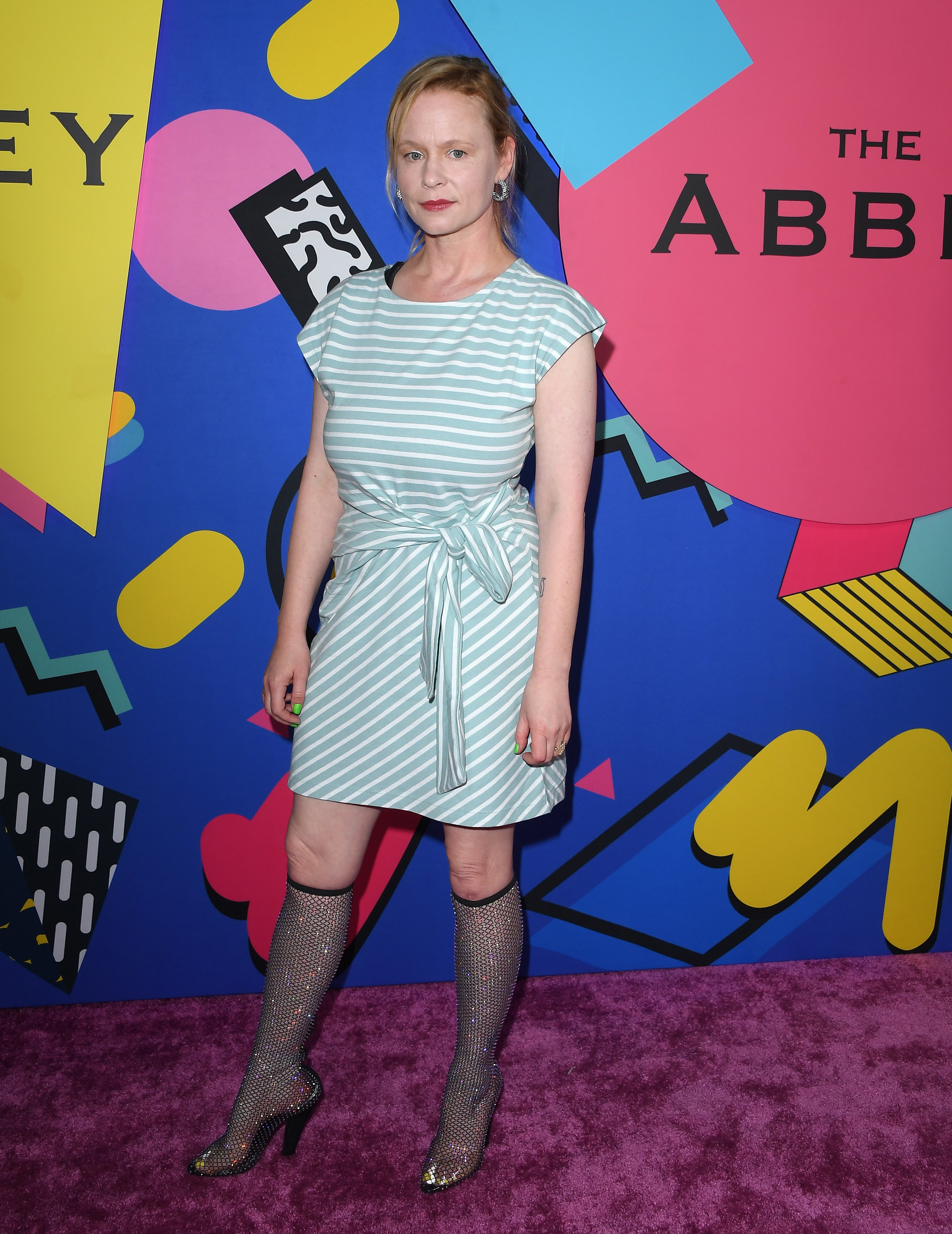 Person in a striped dress and sparkling knee-high boots attends an event in front of a colorful geometric backdrop