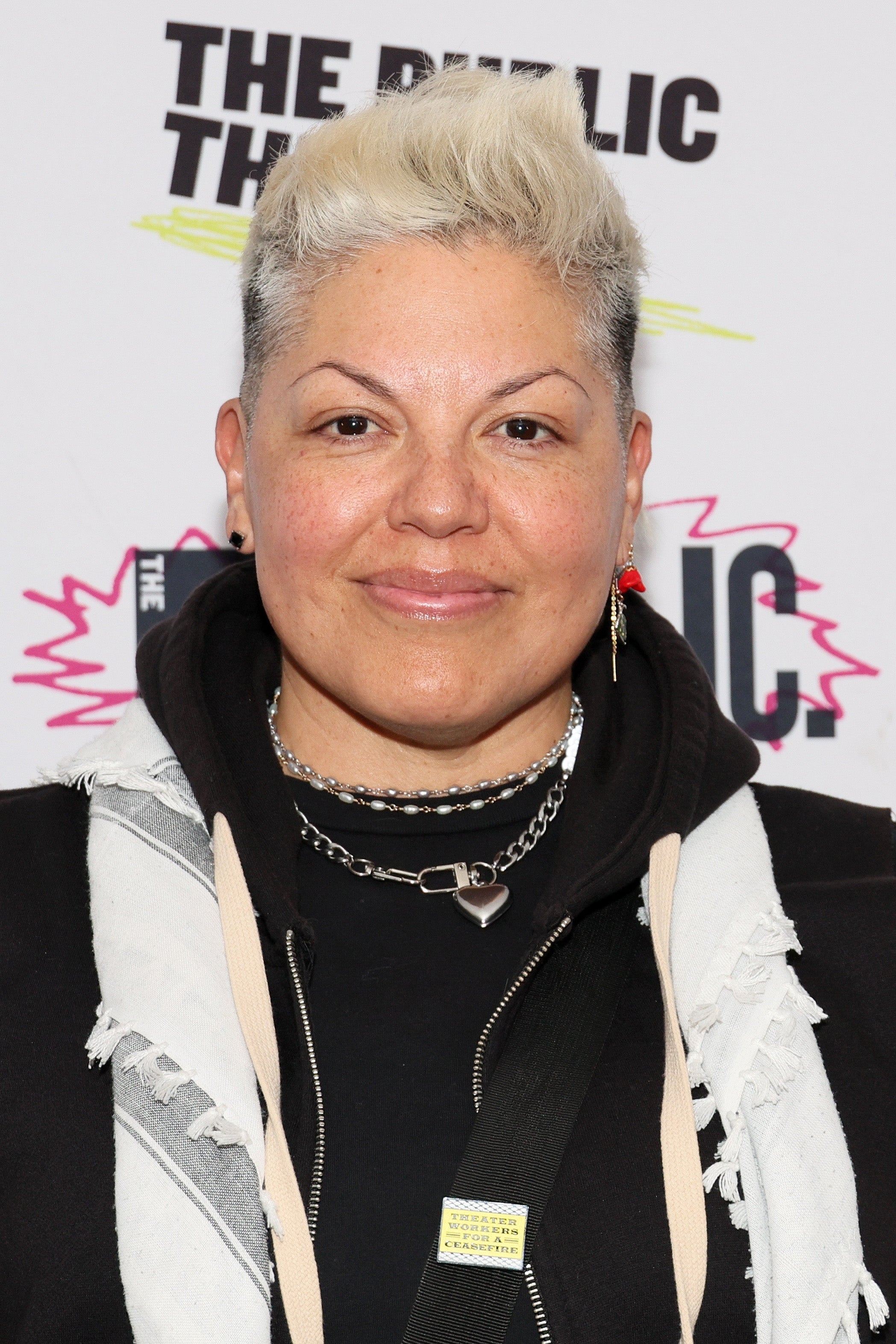 Person with short hair, wearing a casual hoodie and jacket, accessorized with a heart necklace and earrings, at a public event backdrop