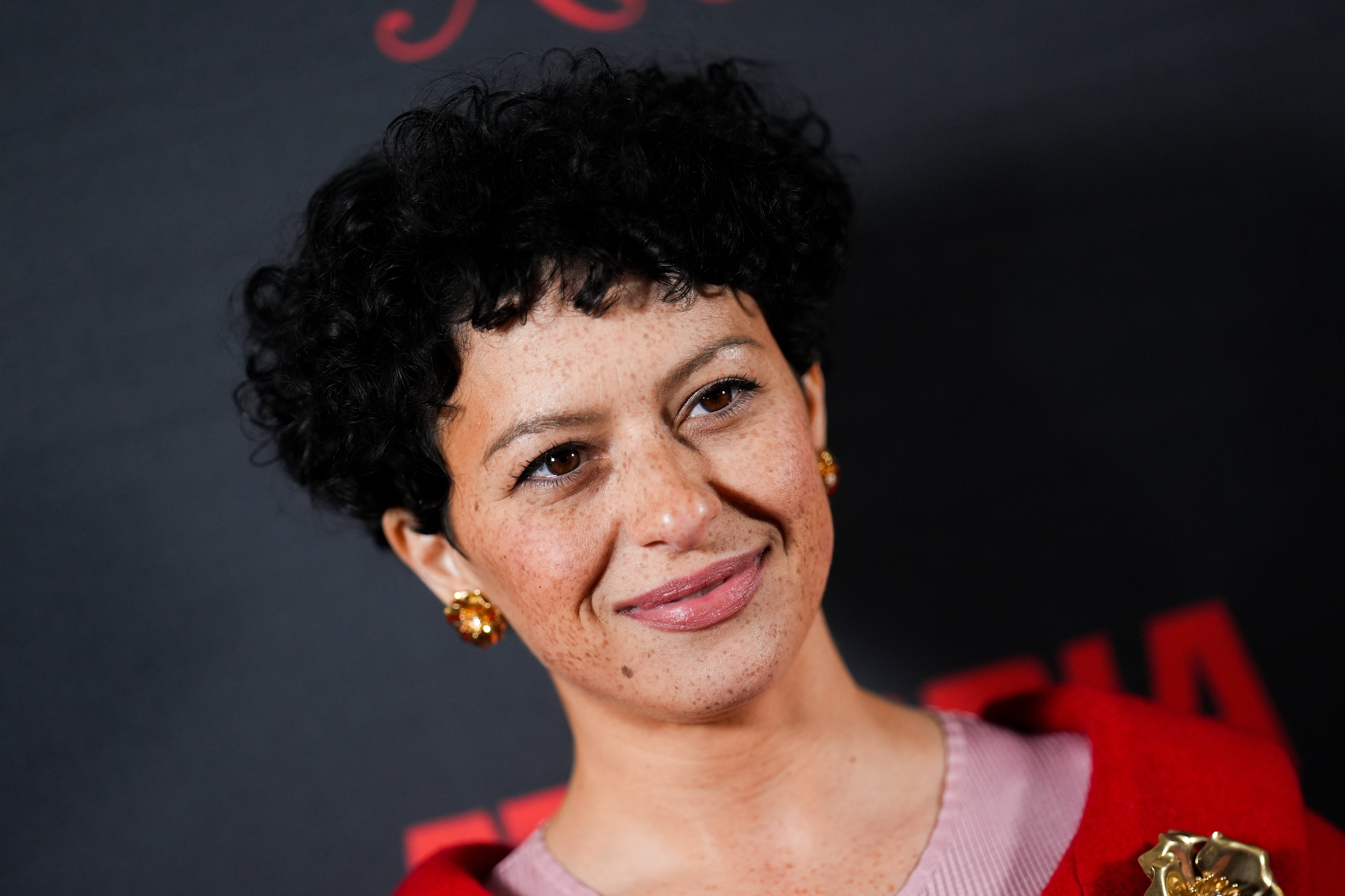 Person with curly hair and freckled skin, wearing a stylish outfit with a brooch, on a red carpet at an event