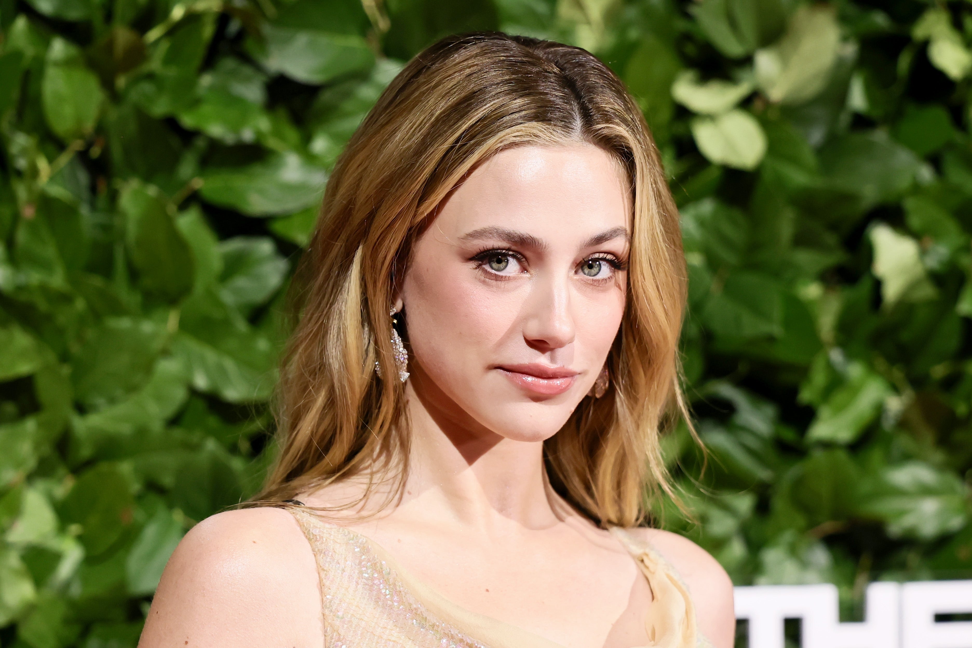 A person on a green foliage backdrop, wearing elegant earrings and a stylish, shimmering dress, looking at the camera