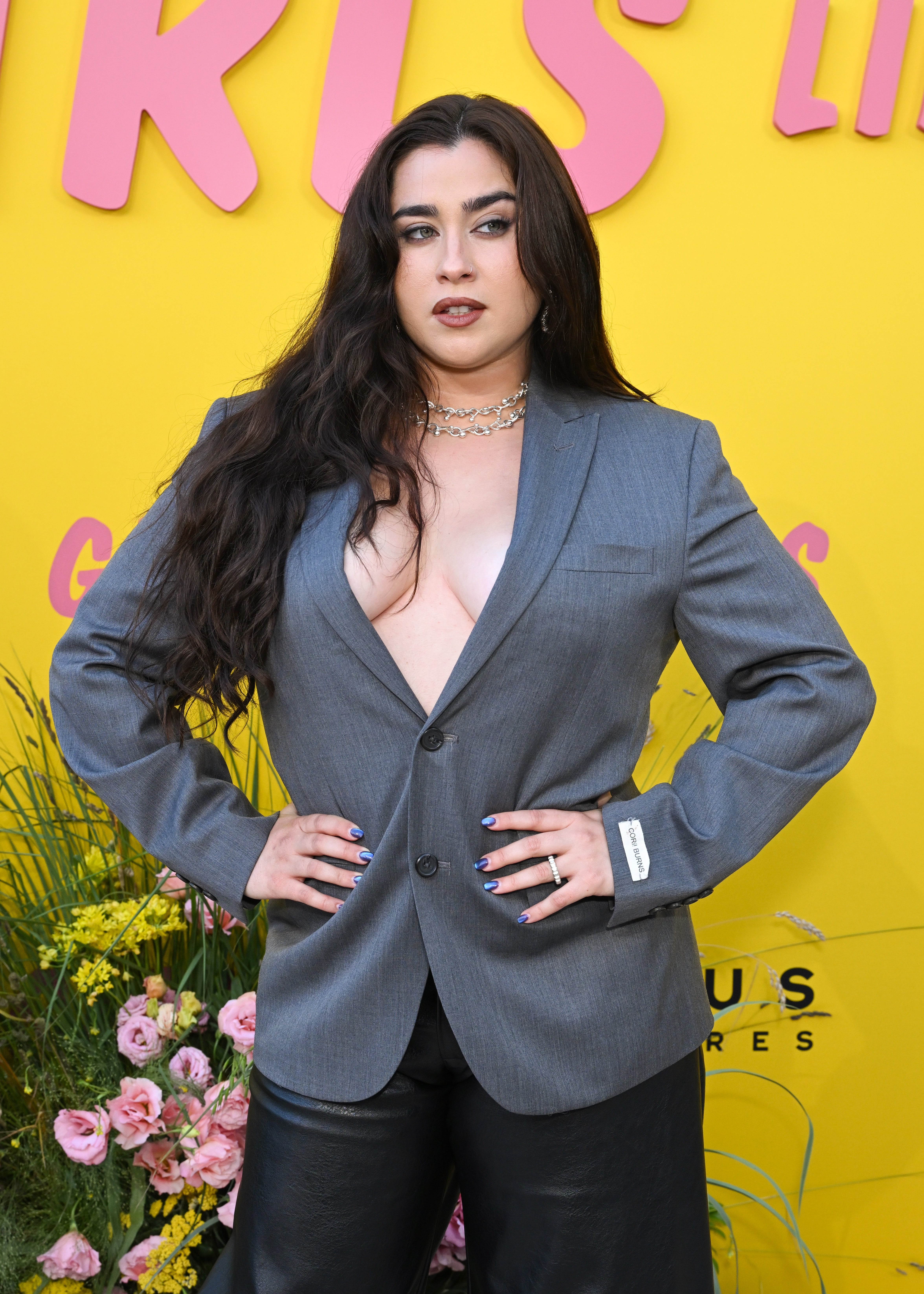 A person poses confidently at an event. They're wearing a stylish open blazer with no shirt, layered necklaces, and black pants