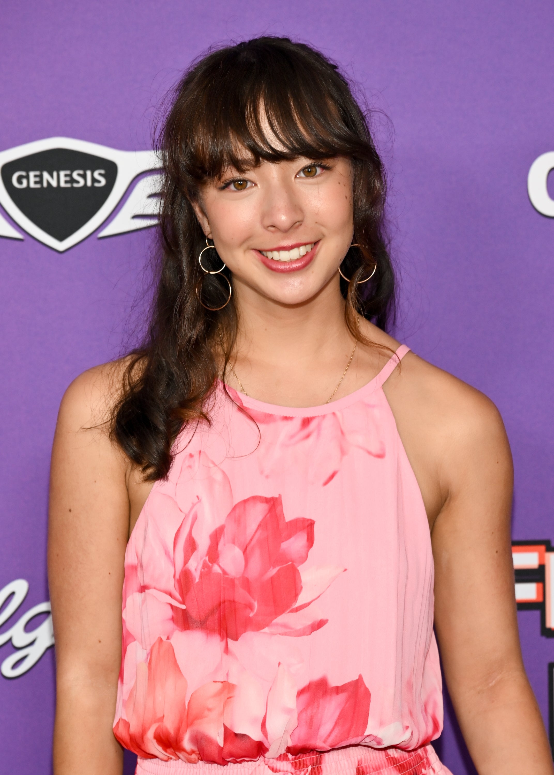 Person in floral-patterned sleeveless dress, attending an event. They are smiling against a purple backdrop with various logo signs