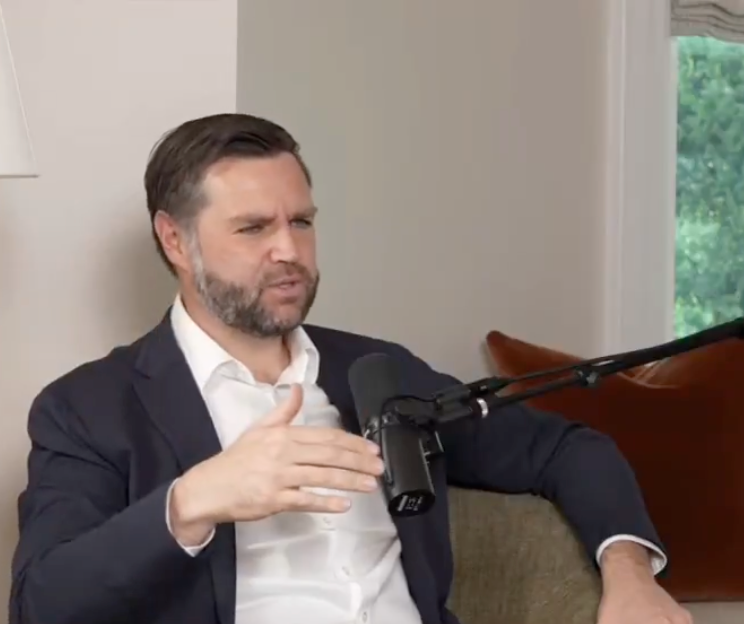 Man in a suit speaking into a microphone during an interview, seated indoors with a neutral background