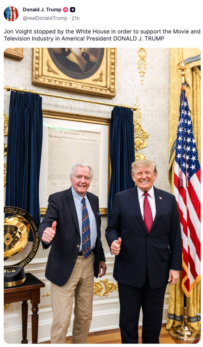Two men stand together in an ornate room, both giving thumbs up. One man wears a suit, the other a patterned blazer with a tie
