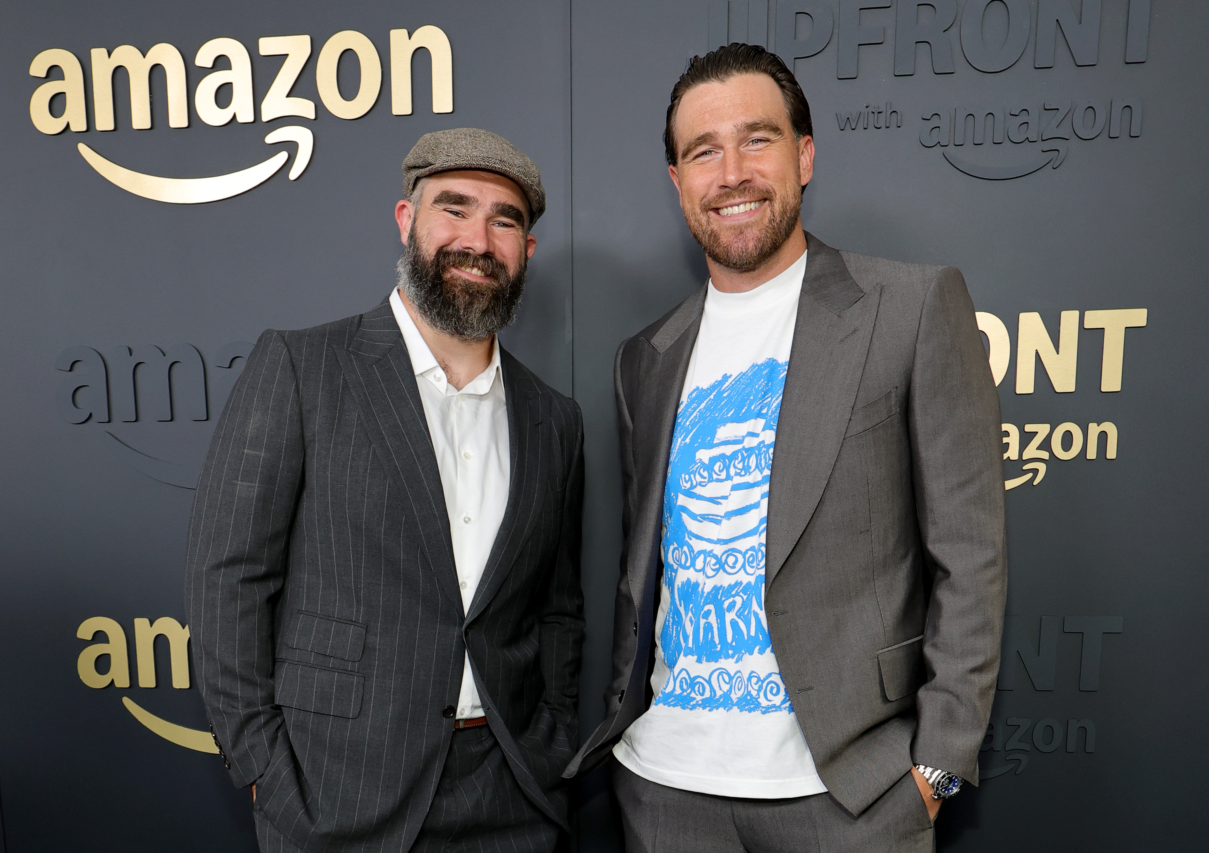 Two men in suits pose on an Amazon event red carpet. One wears a hat; the other sports a printed T-shirt under his suit jacket