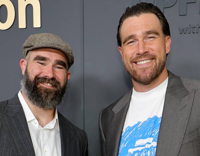 Two men in suits pose on an Amazon event red carpet. One wears a hat; the other sports a printed T-shirt under his suit jacket