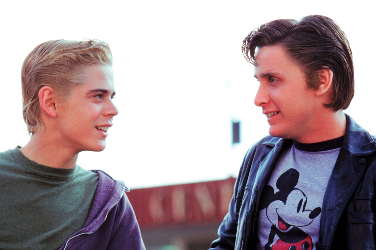 Two young men are smiling at each other outdoors. One wears a jacket over a Mickey Mouse shirt, the other wears a hoodie over a T-shirt