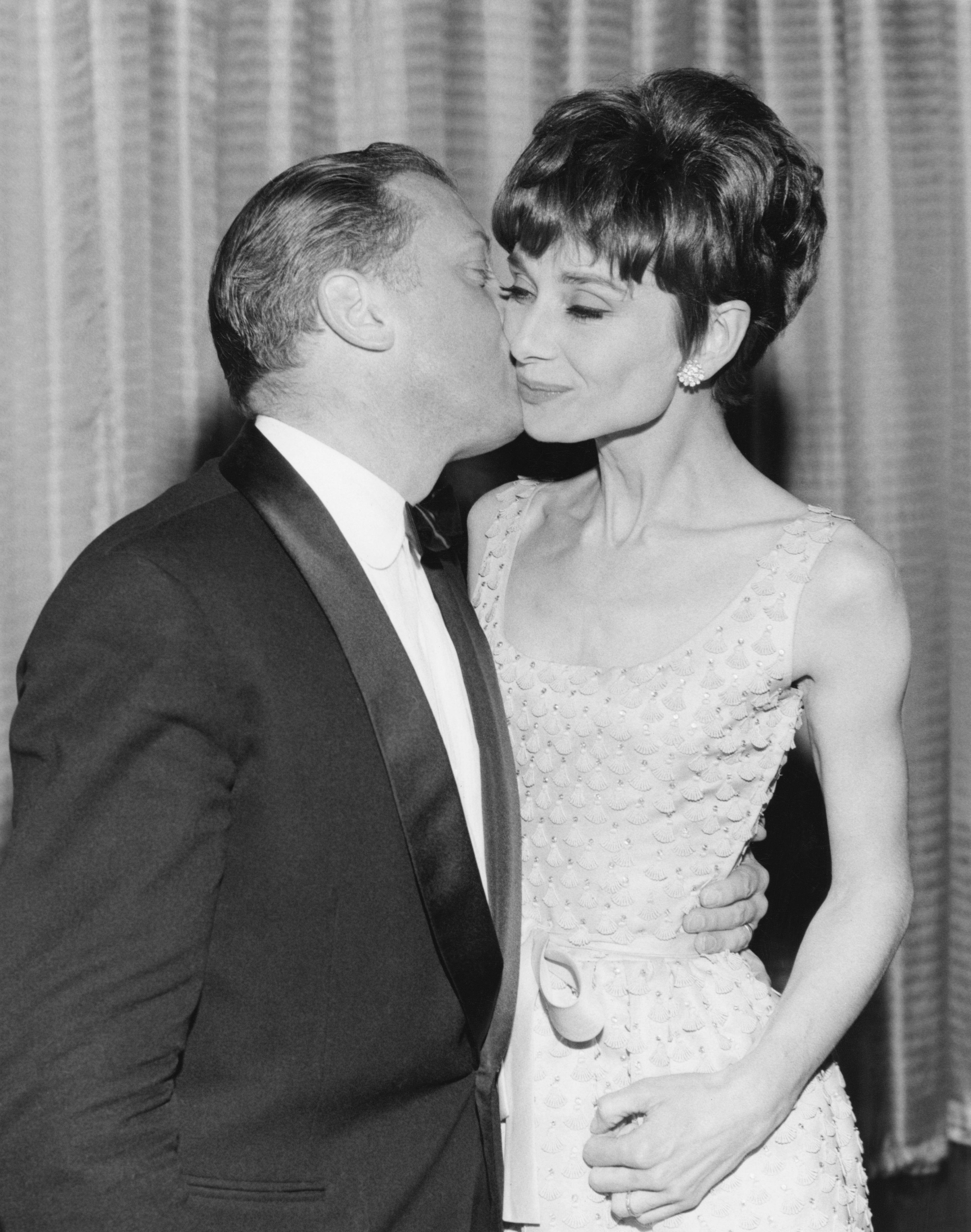 Richard Attenborough in a tuxedo kisses a smiling Audrey Hepburn in a sleeveless, textured gown at an event