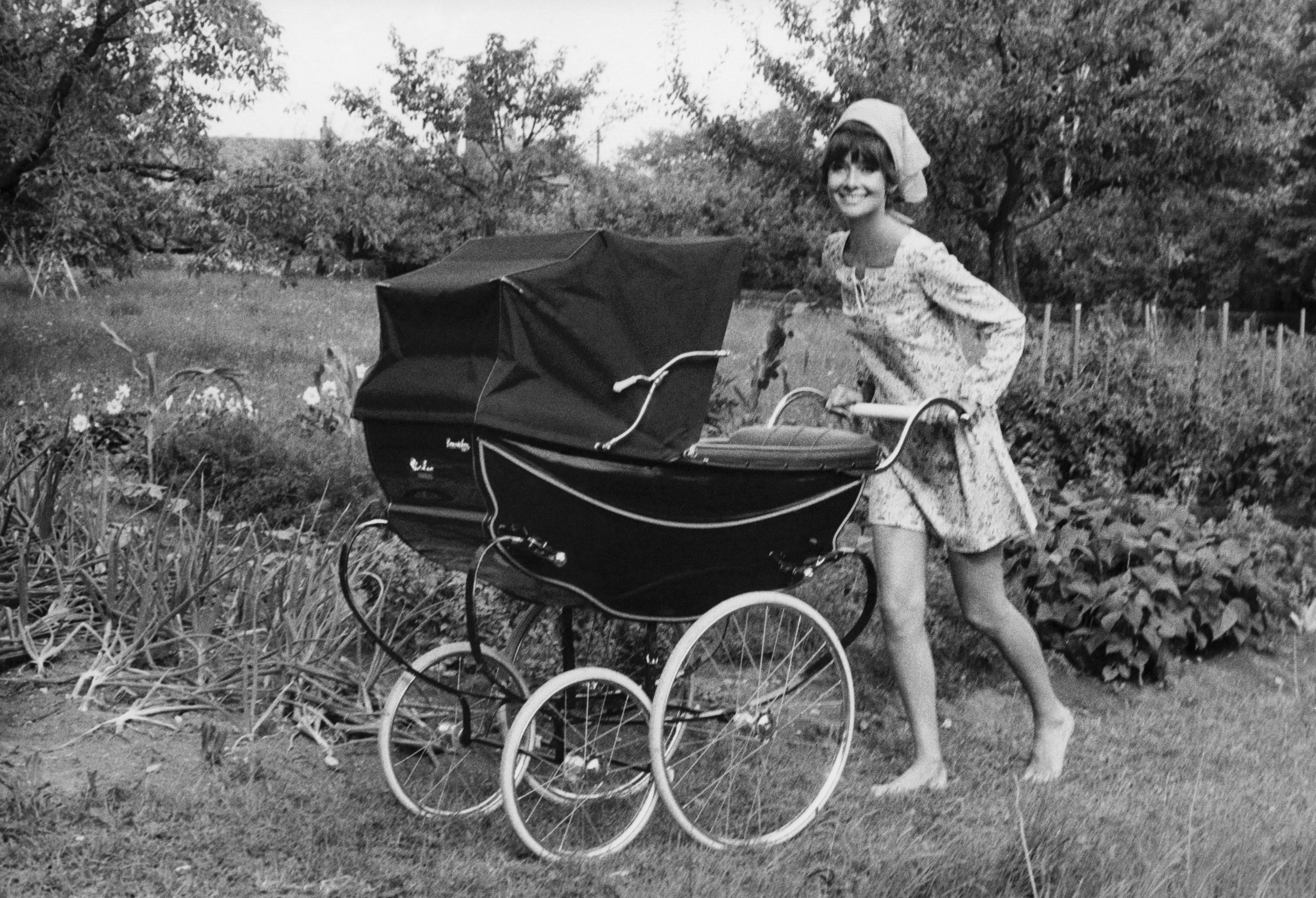 Audrey Hepburn in a vintage dress and headscarf smiles while playfully pushing a classic pram in a garden setting