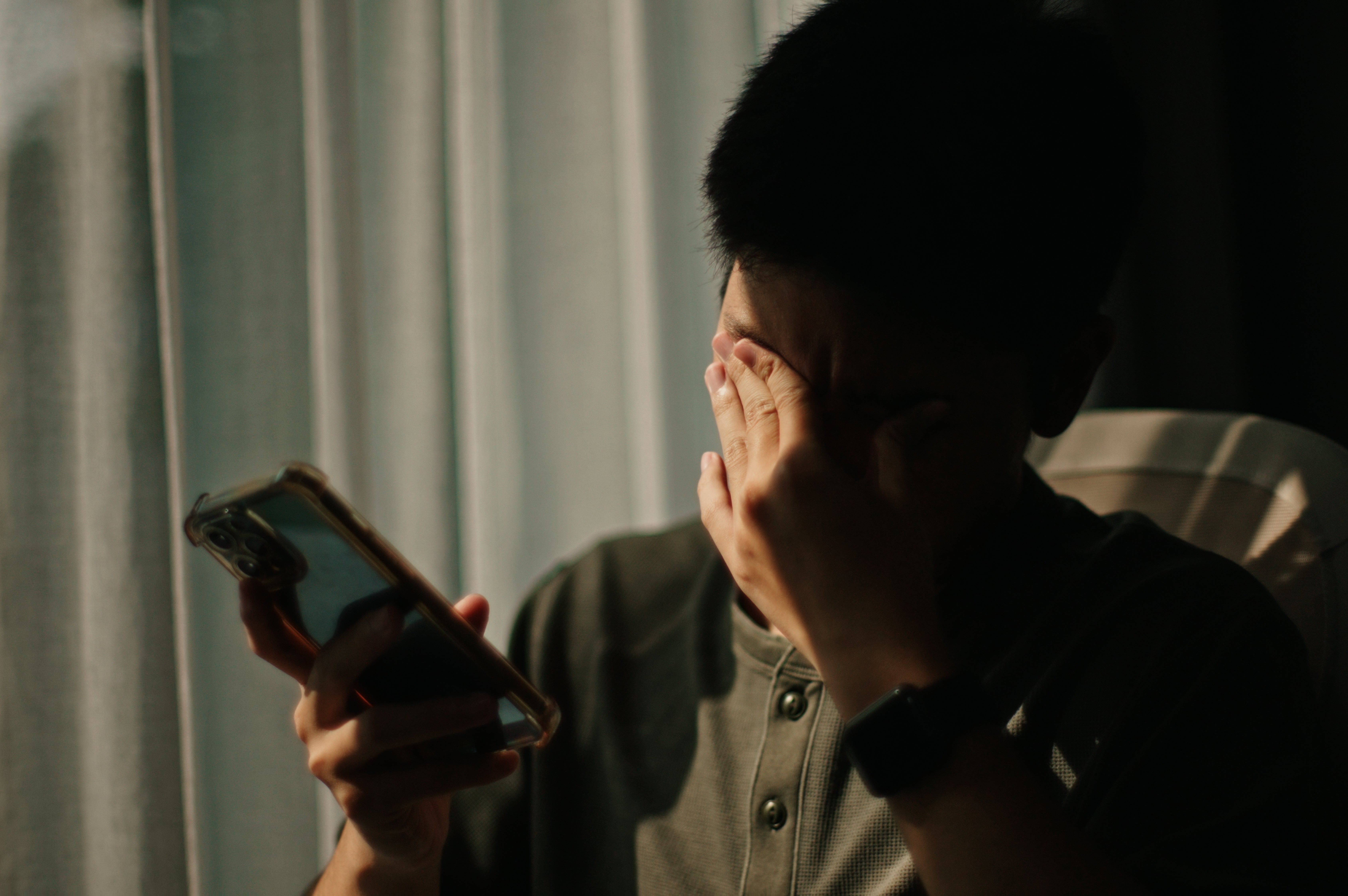 Person sitting by curtains, covering face with one hand, holding a smartphone in the other, suggesting distress or deep thought