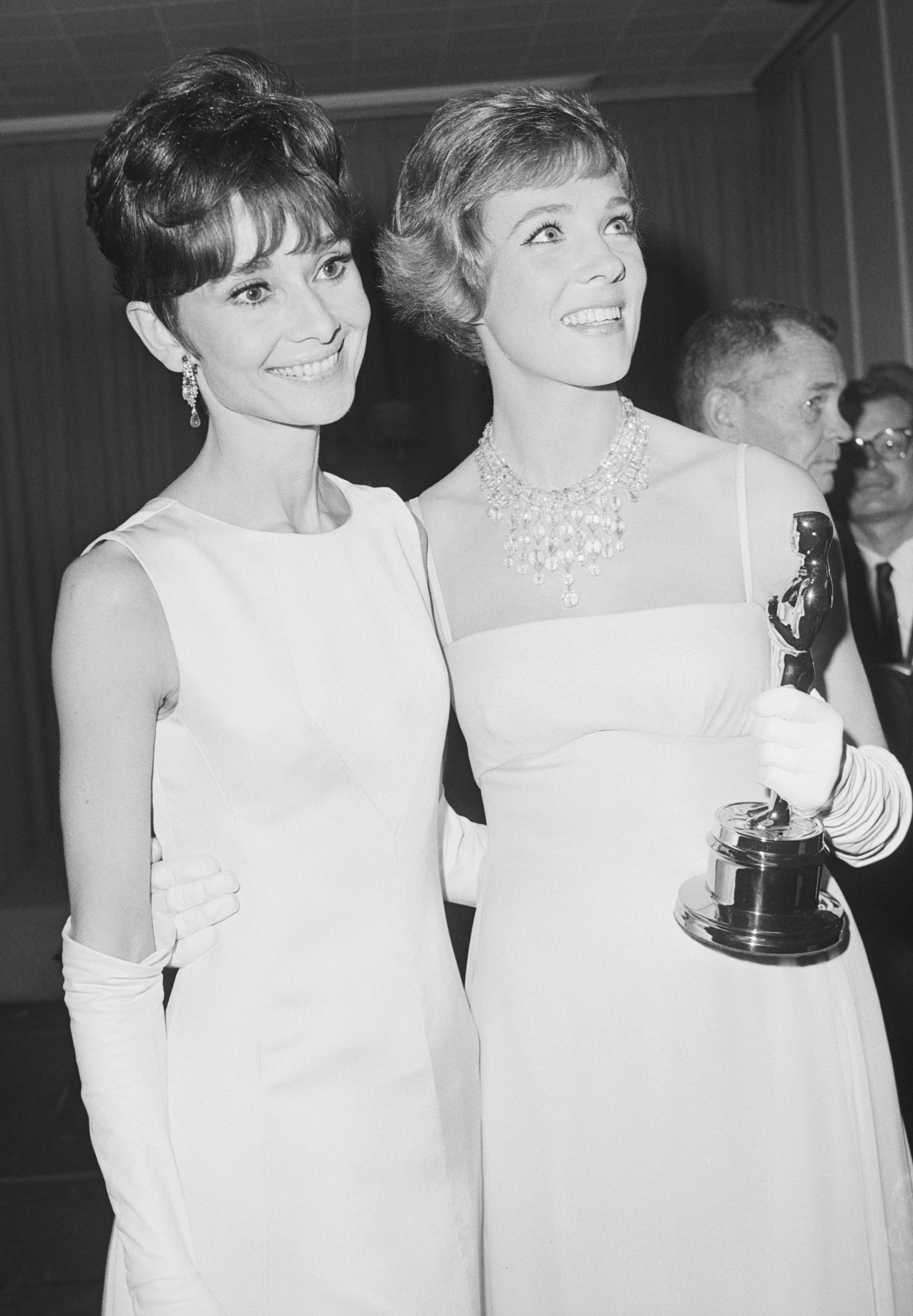 Audrey and Julie in elegant gowns and gloves, one holding an award statue, standing and smiling at an event