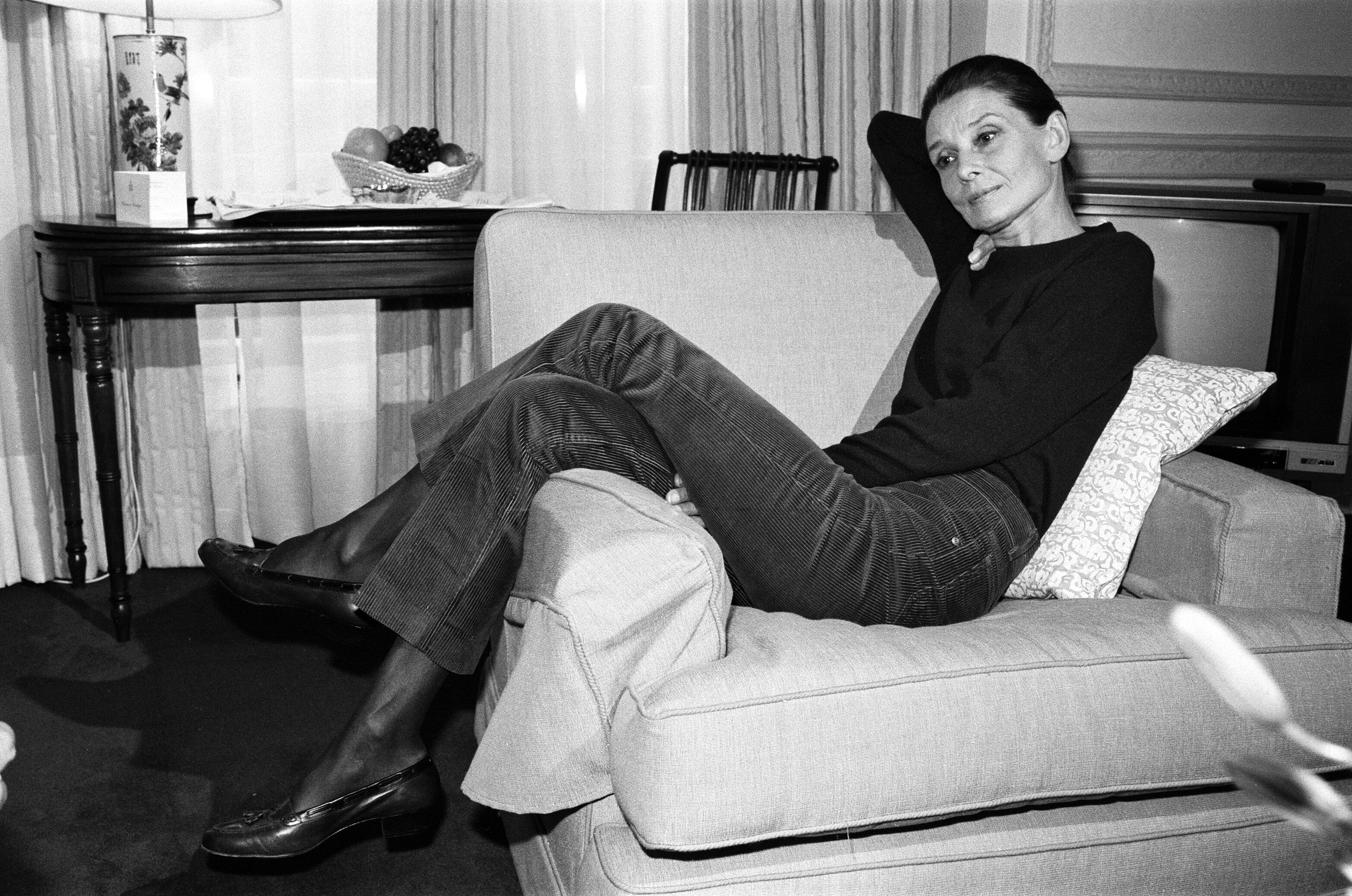 Audrey Hepburn lounges on a sofa in a relaxed pose, wearing a sweater and corduroy pants. A lamp and a table with a book are in the background