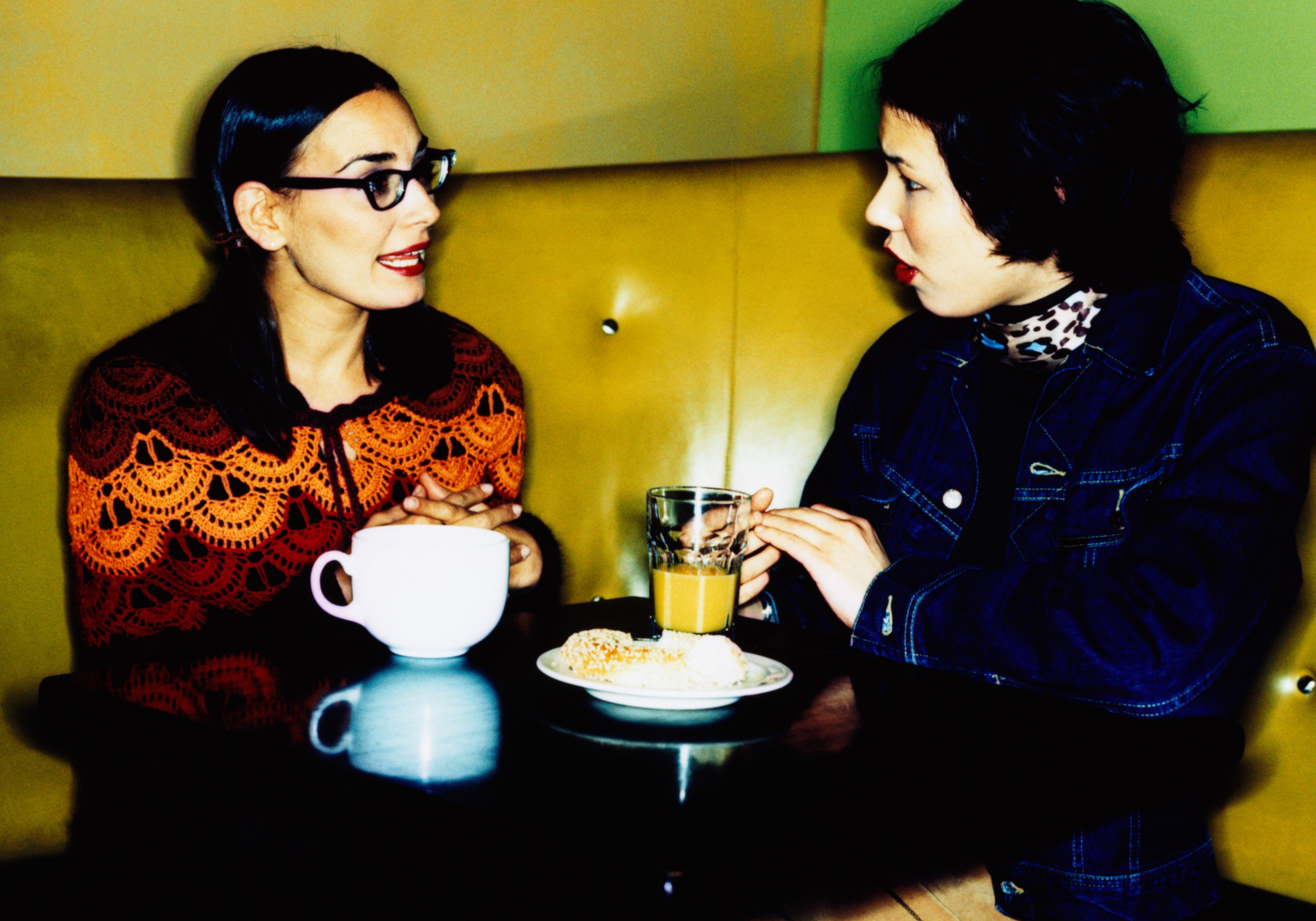 Two people sit at a cafe table engaged in conversation, with drinks and snacks in front of them