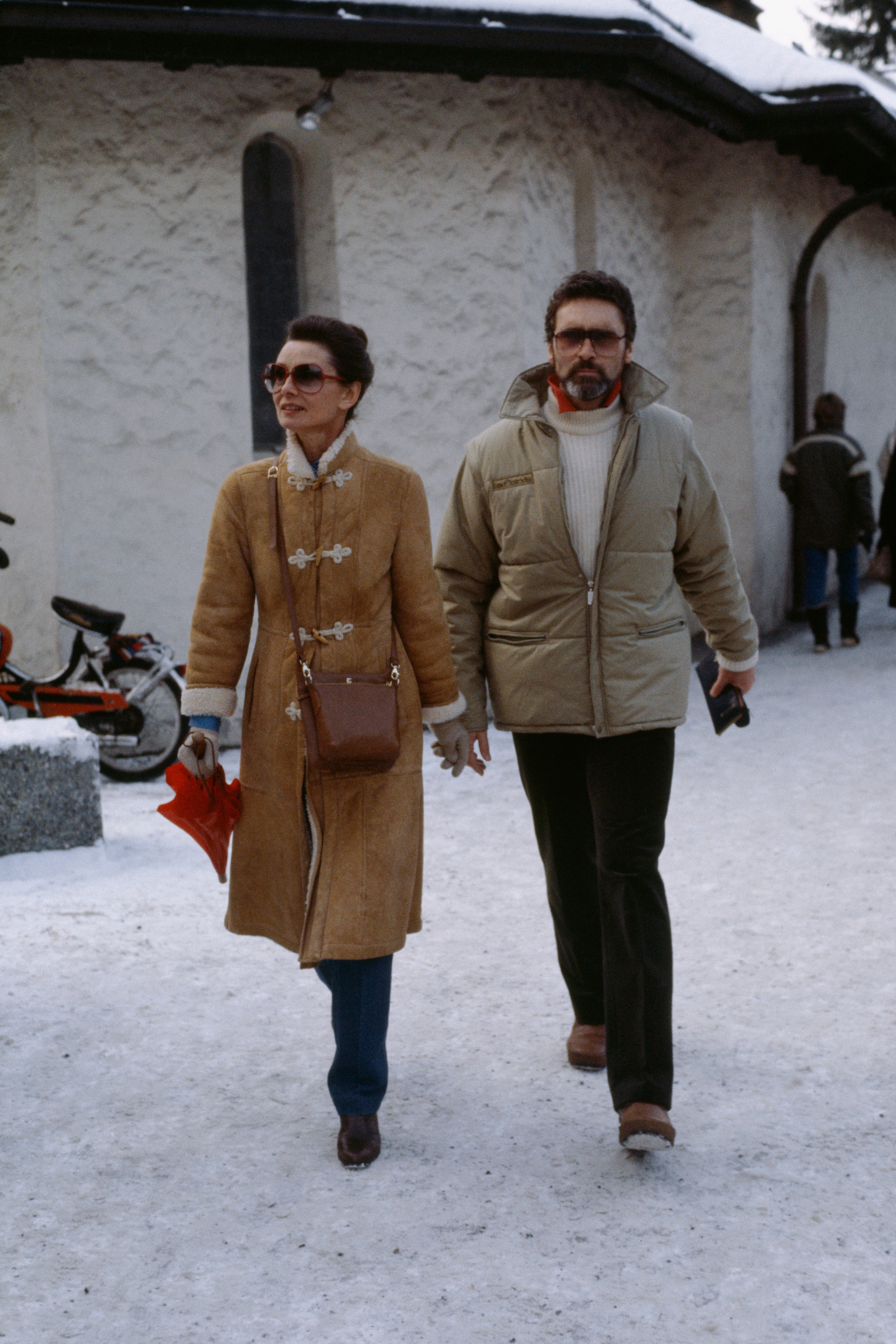 A couple walks hand in hand on snow. Audrey wears a stylish long coat with flower details; Robert wears a cozy jacket and turtleneck