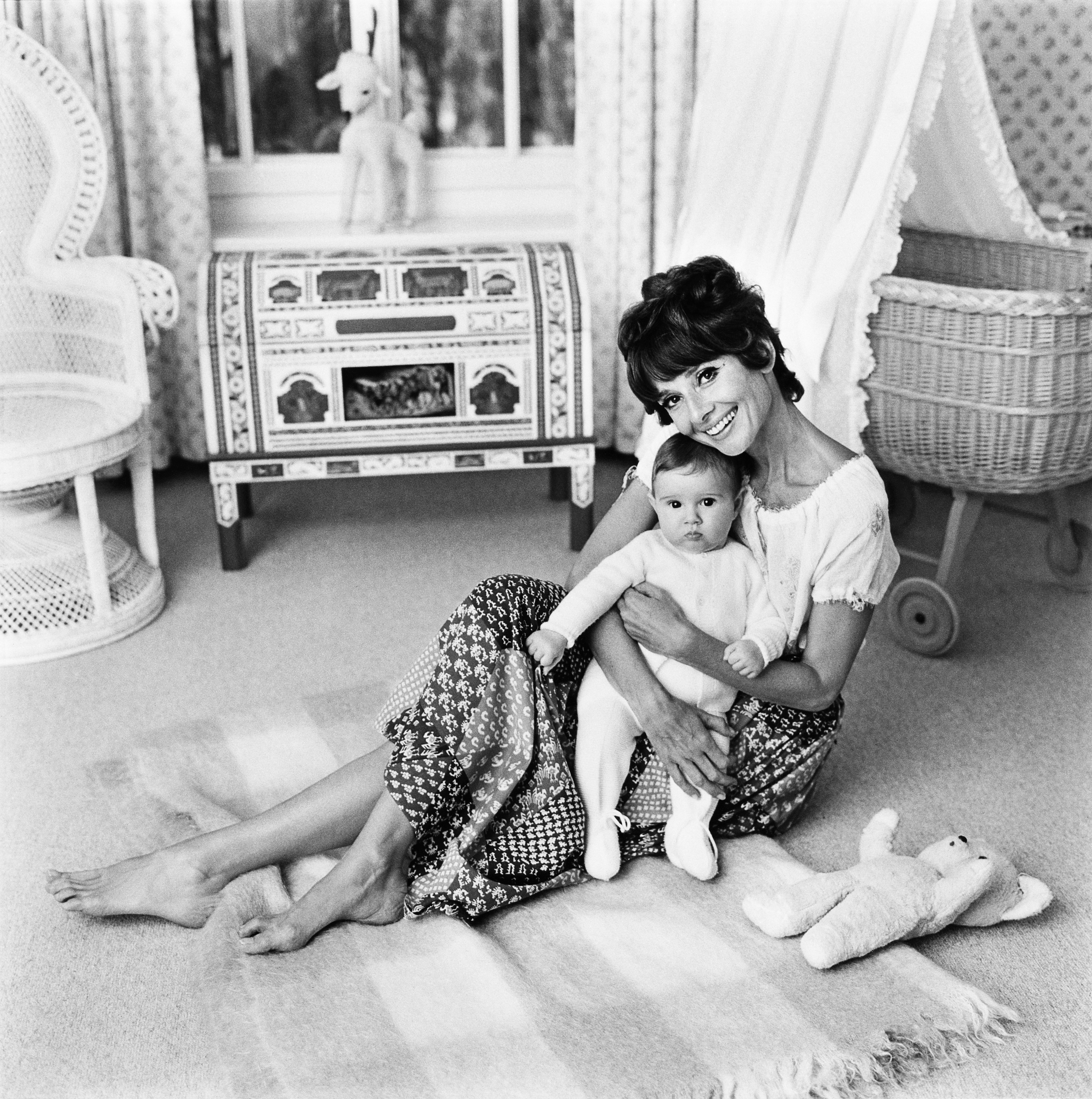 Audrey Hepburn in a bohemian-style skirt sits on a rug, holding a baby. They're in a stylishly decorated room with toys and a wicker chair