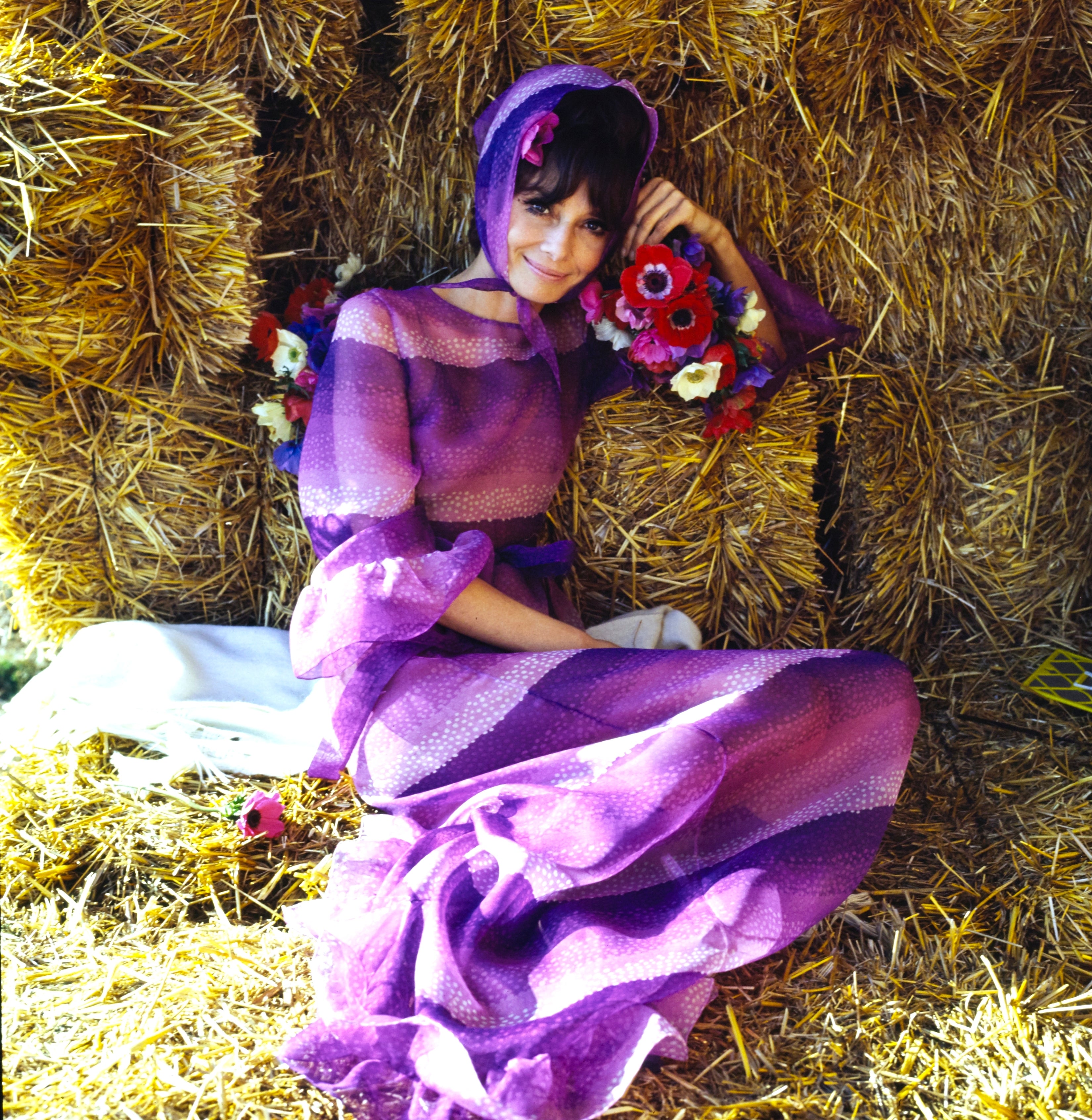 Audrey Hepburn in a flowing dress with a headscarf, sitting on hay bales holding colorful flowers