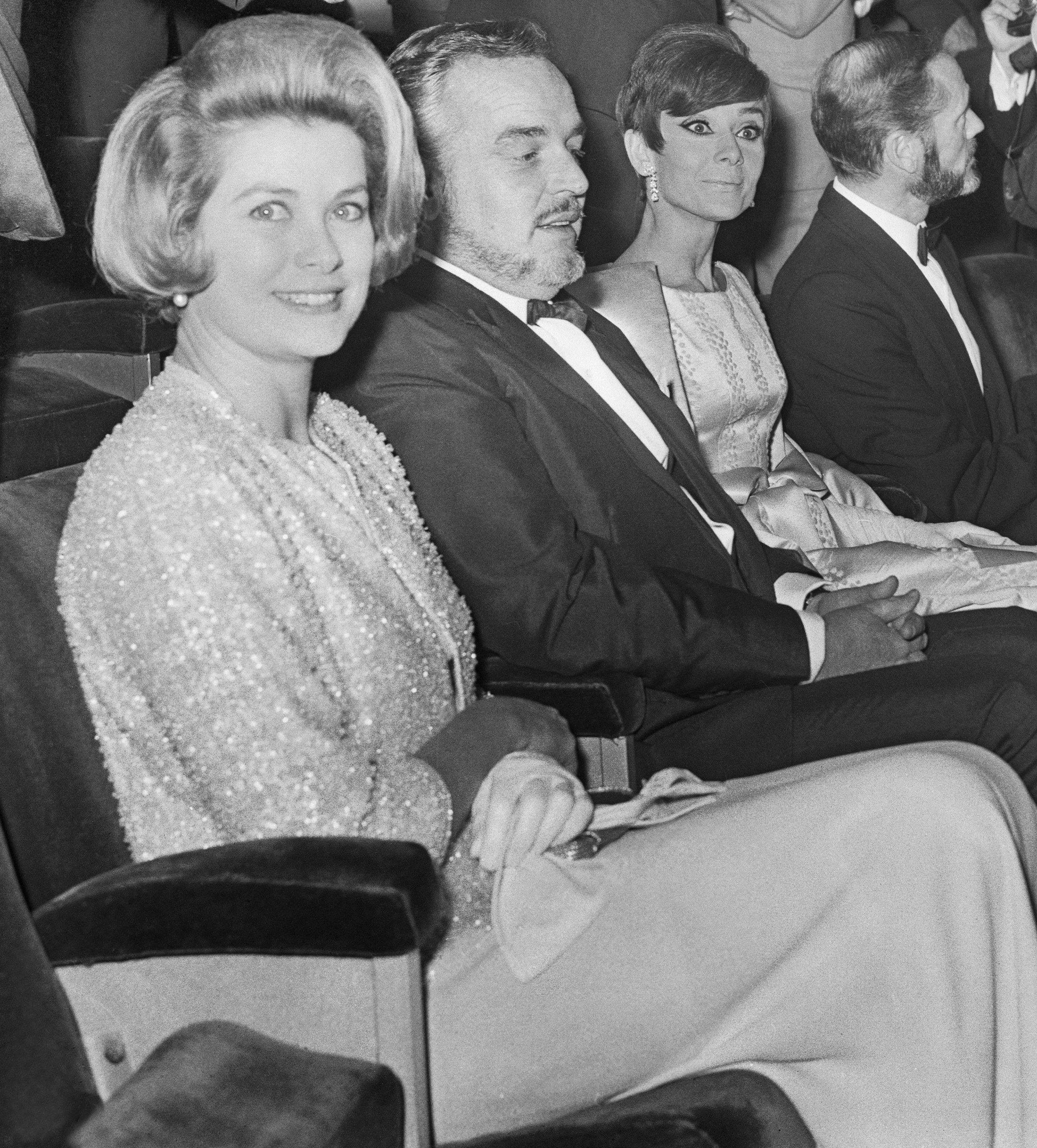 Celebrities seated at an event, wearing formal attire; photographers and audience in background capturing photos