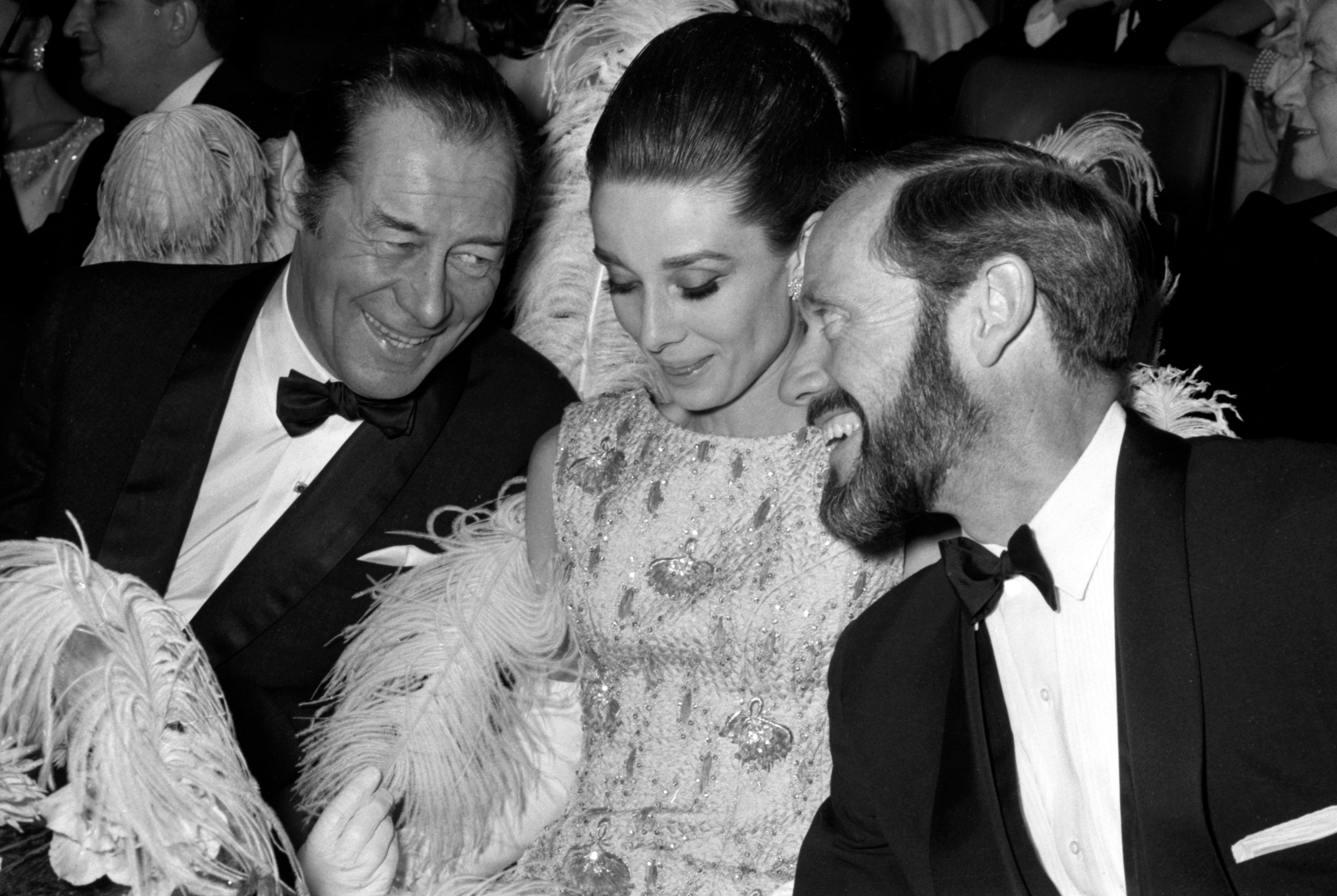 A glamorous event with three people dressed in elegant evening wear adorned with feathers, engaged in conversation and smiling