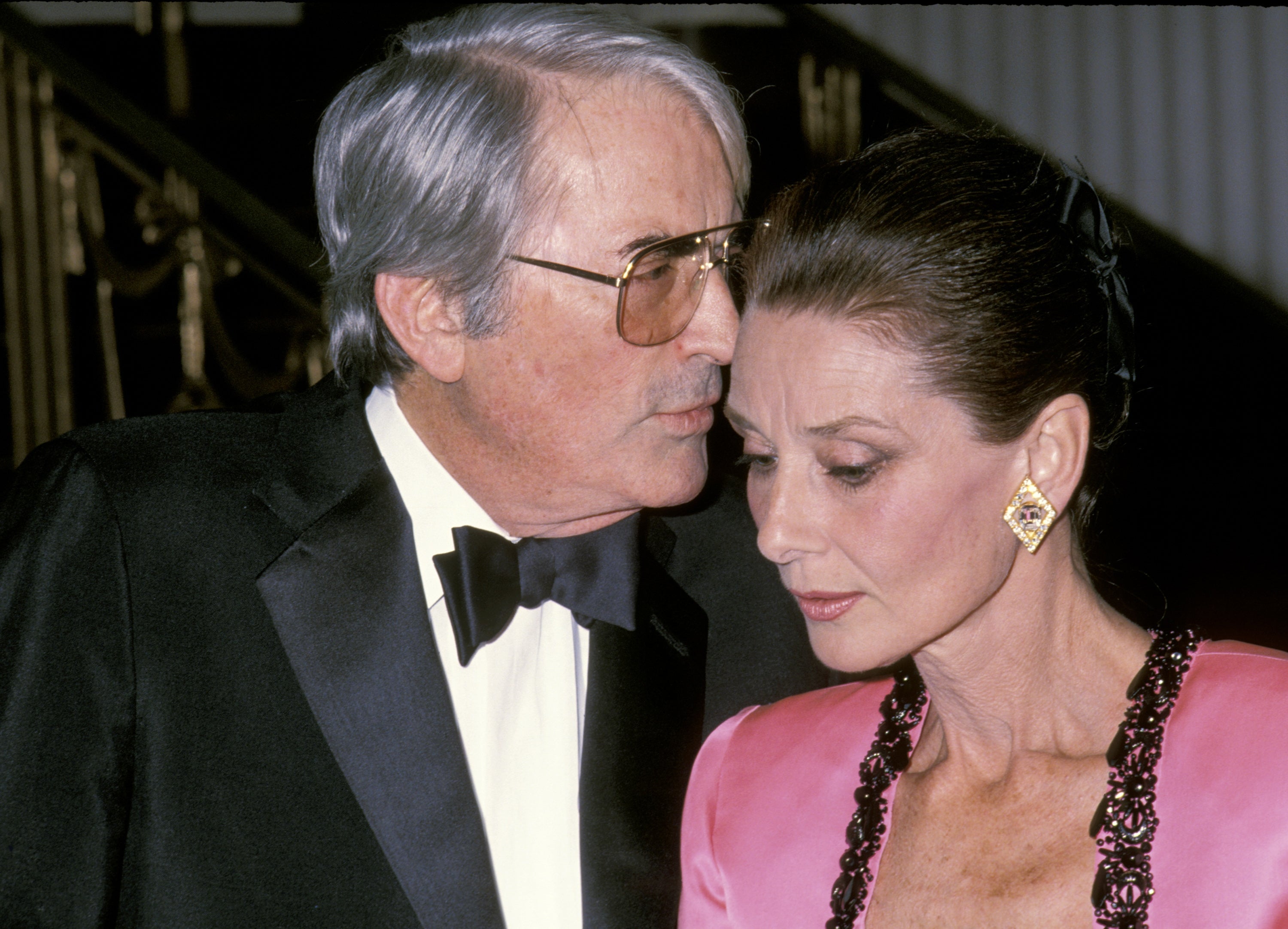 Gregory in a tuxedo whispers to Audrey in a pink, elegant gown with statement earrings at a formal event