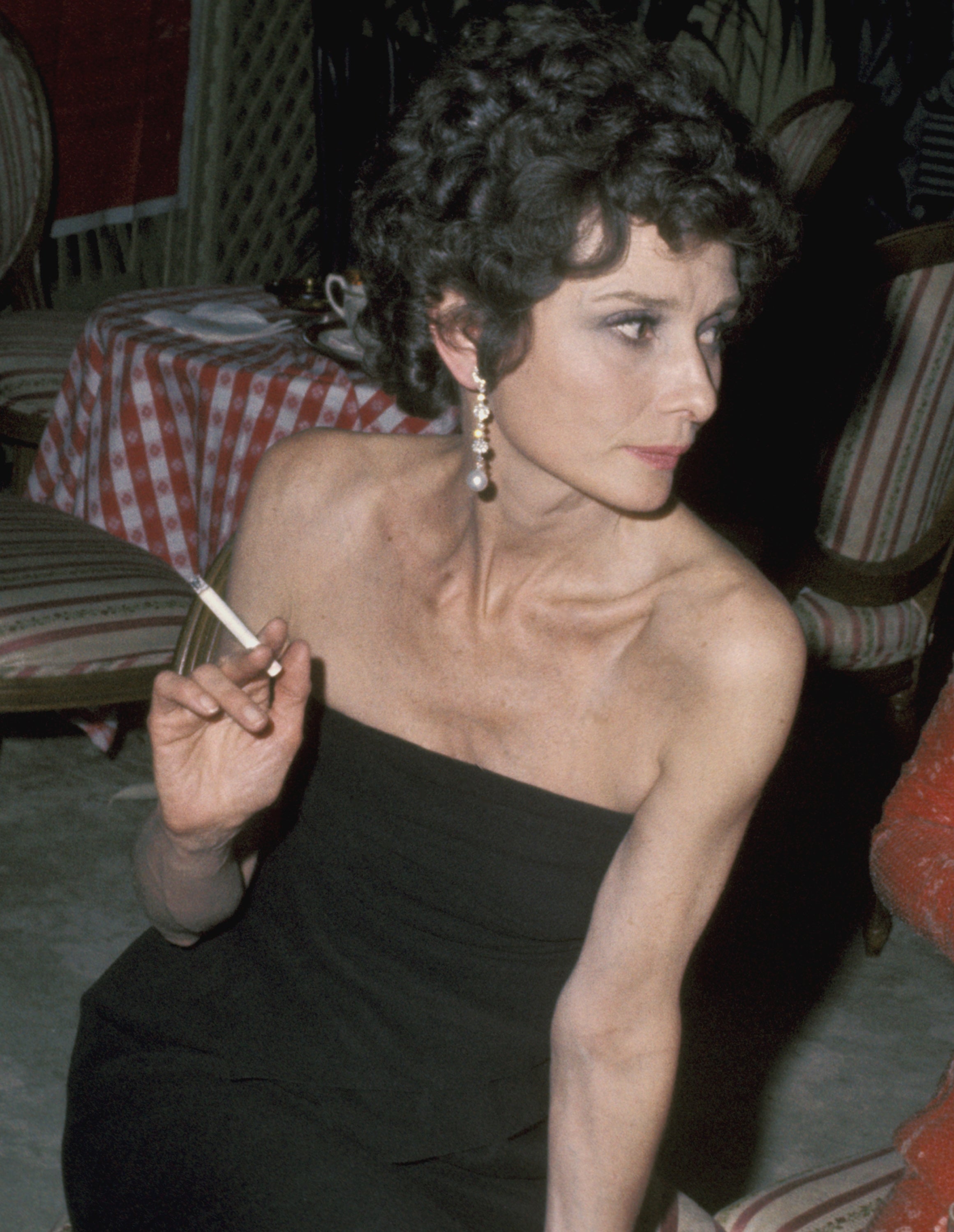 Audrey Hepburn seated in a restaurant, wearing a strapless dress and dangling earrings, holding a cigarette