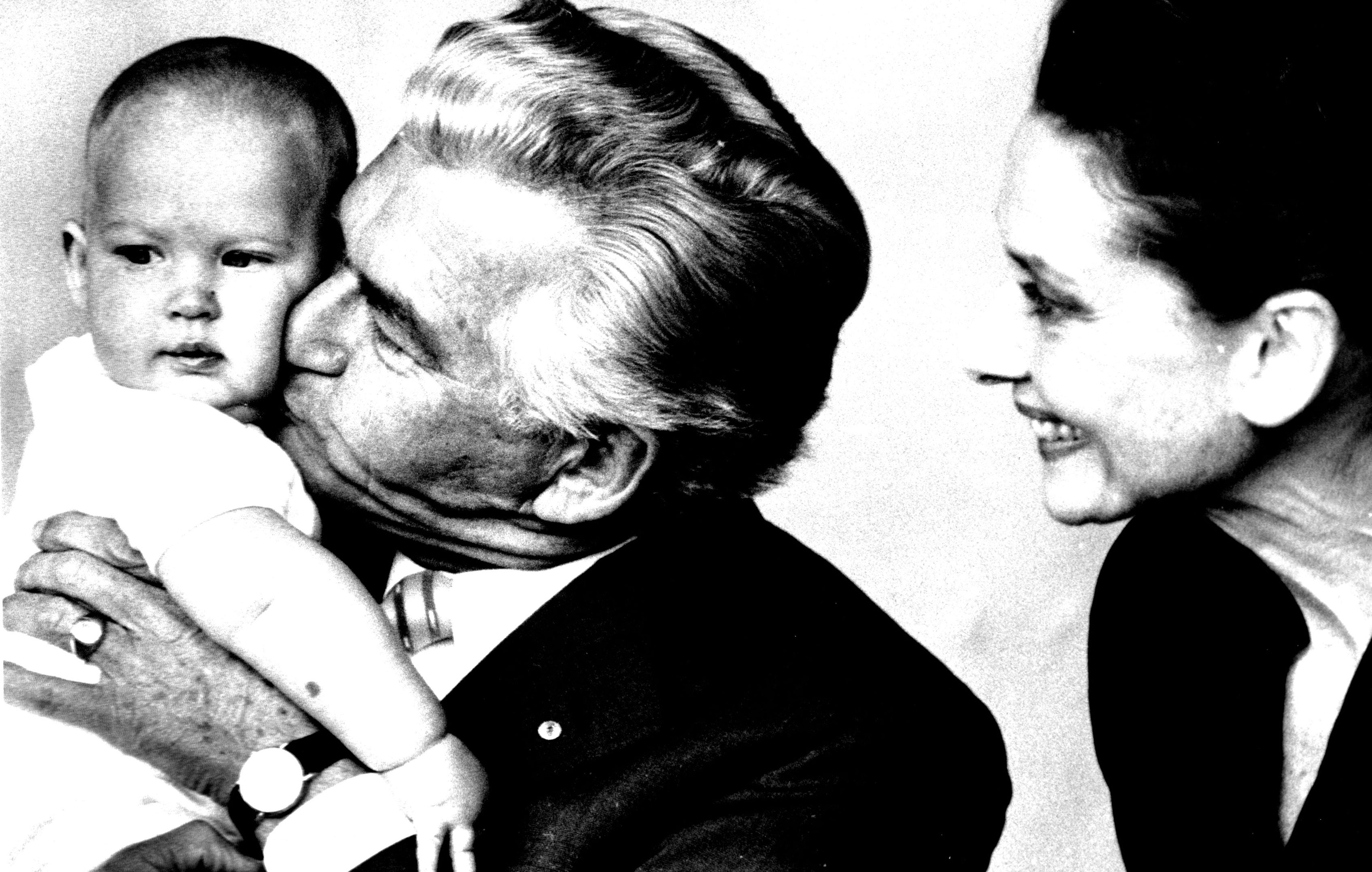 Bob Hawke kisses a baby while a smiling Audrey Hepburn woman looks on. They appear joyous and affectionate