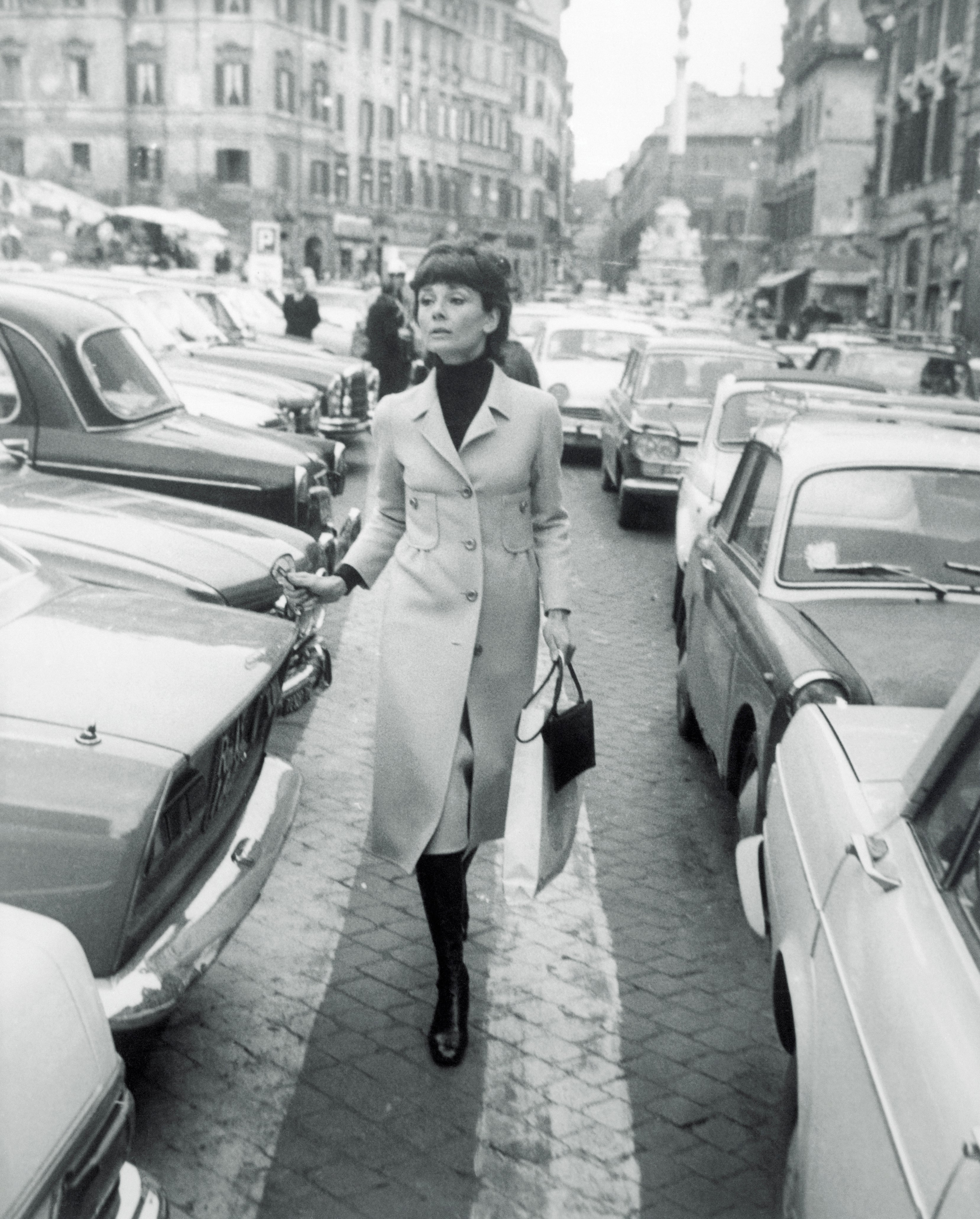 Audrey Hepburn wearing a long coat and boots, holding a bag, walking confidently between parked cars on a city street