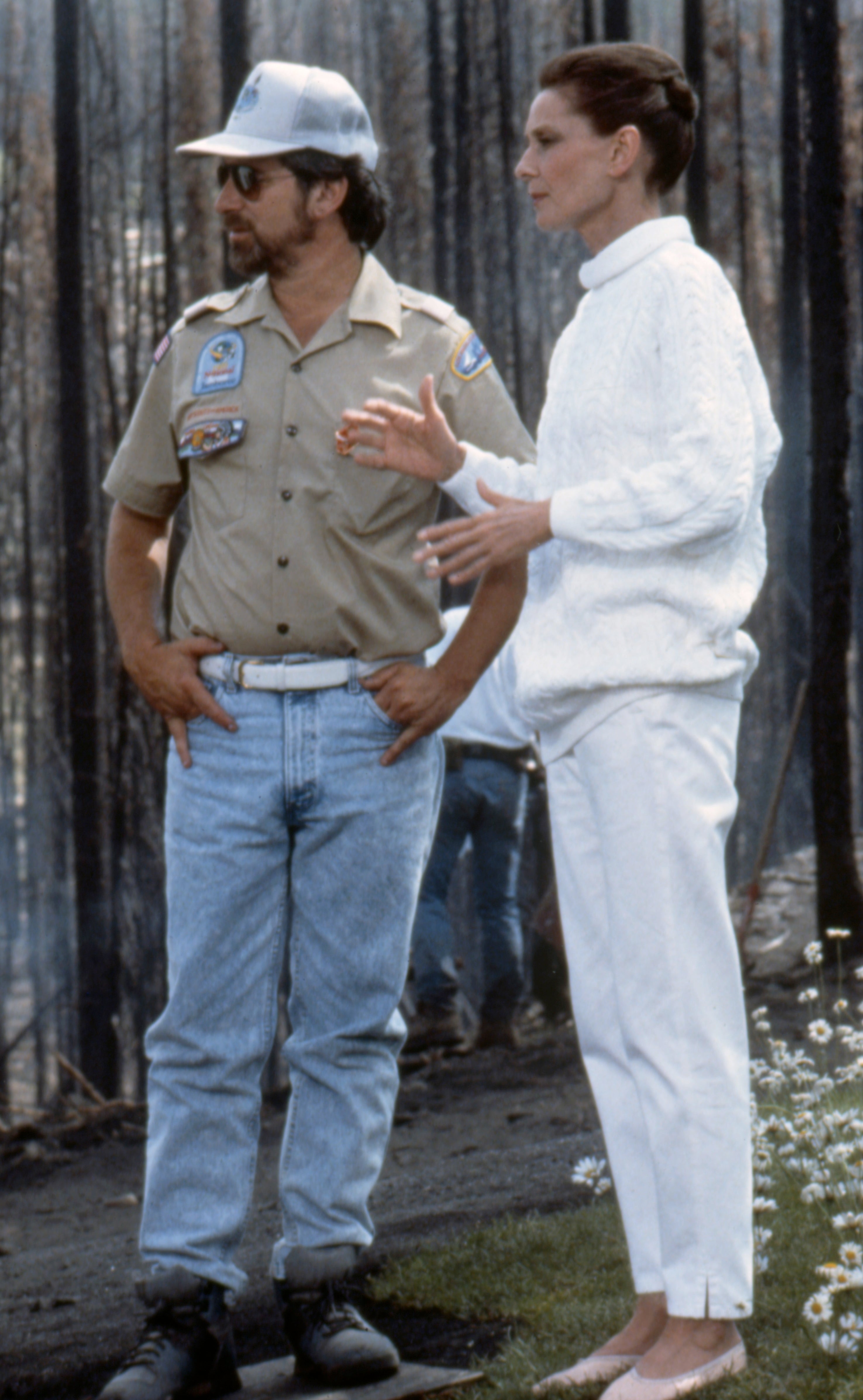 Audrey Hepburn in a white sweater and pants talks to Steven Spielberg, standing outdoors near a charred forest area