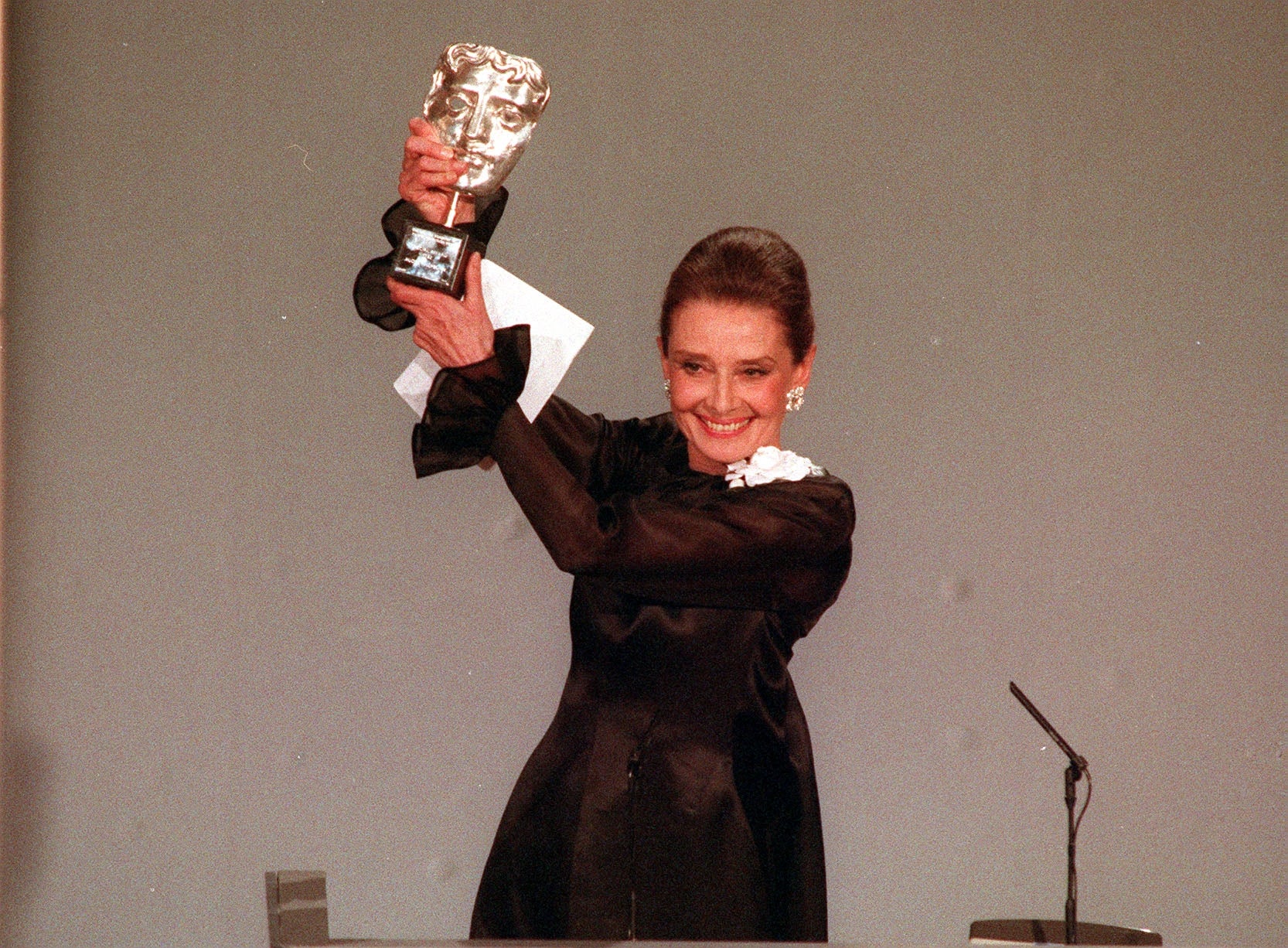 Audrey Hepburn holding a BAFTA award, smiling, in elegant long-sleeve attire with floral detail