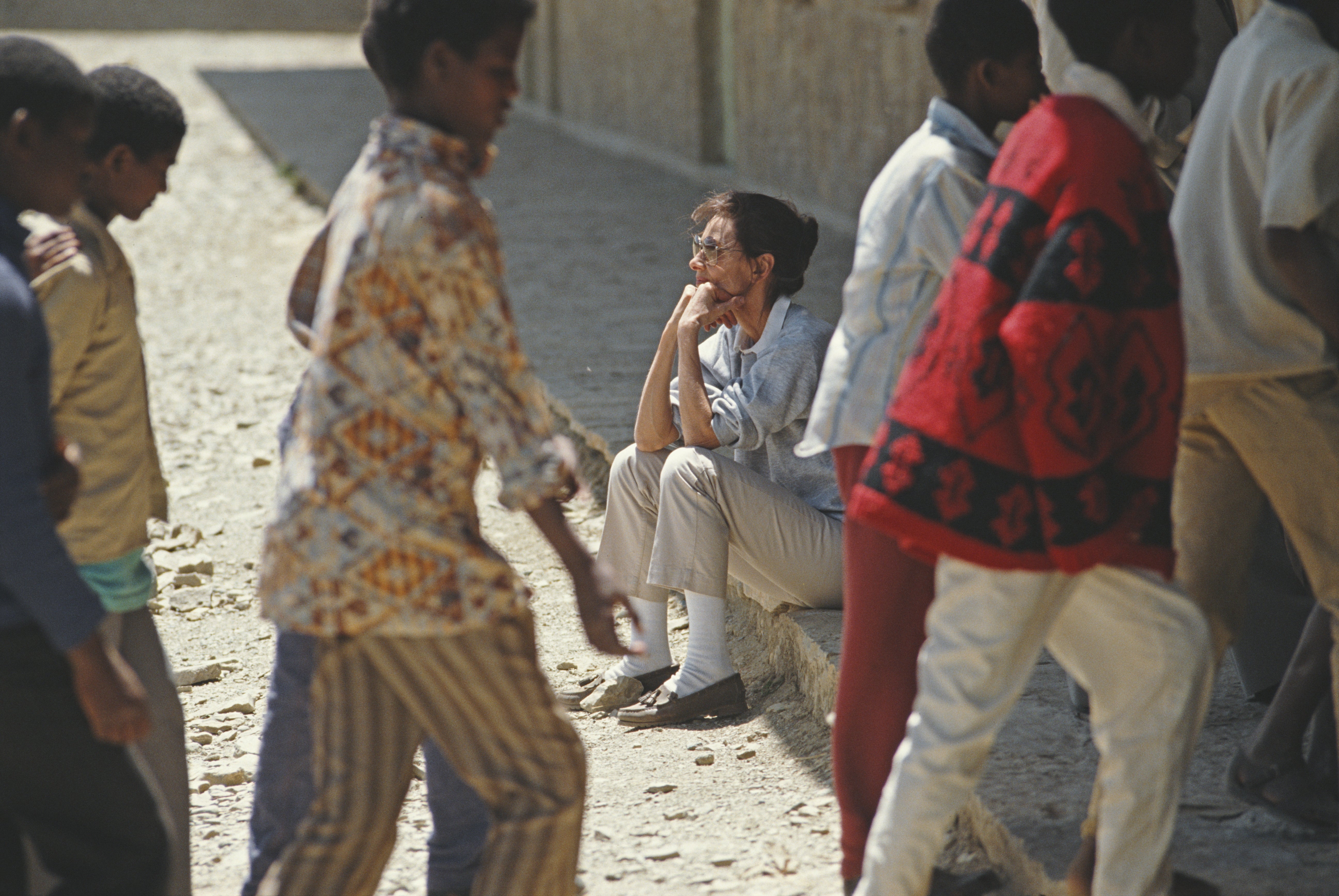 Audrey Hepburn sits on a curb with people walking past her