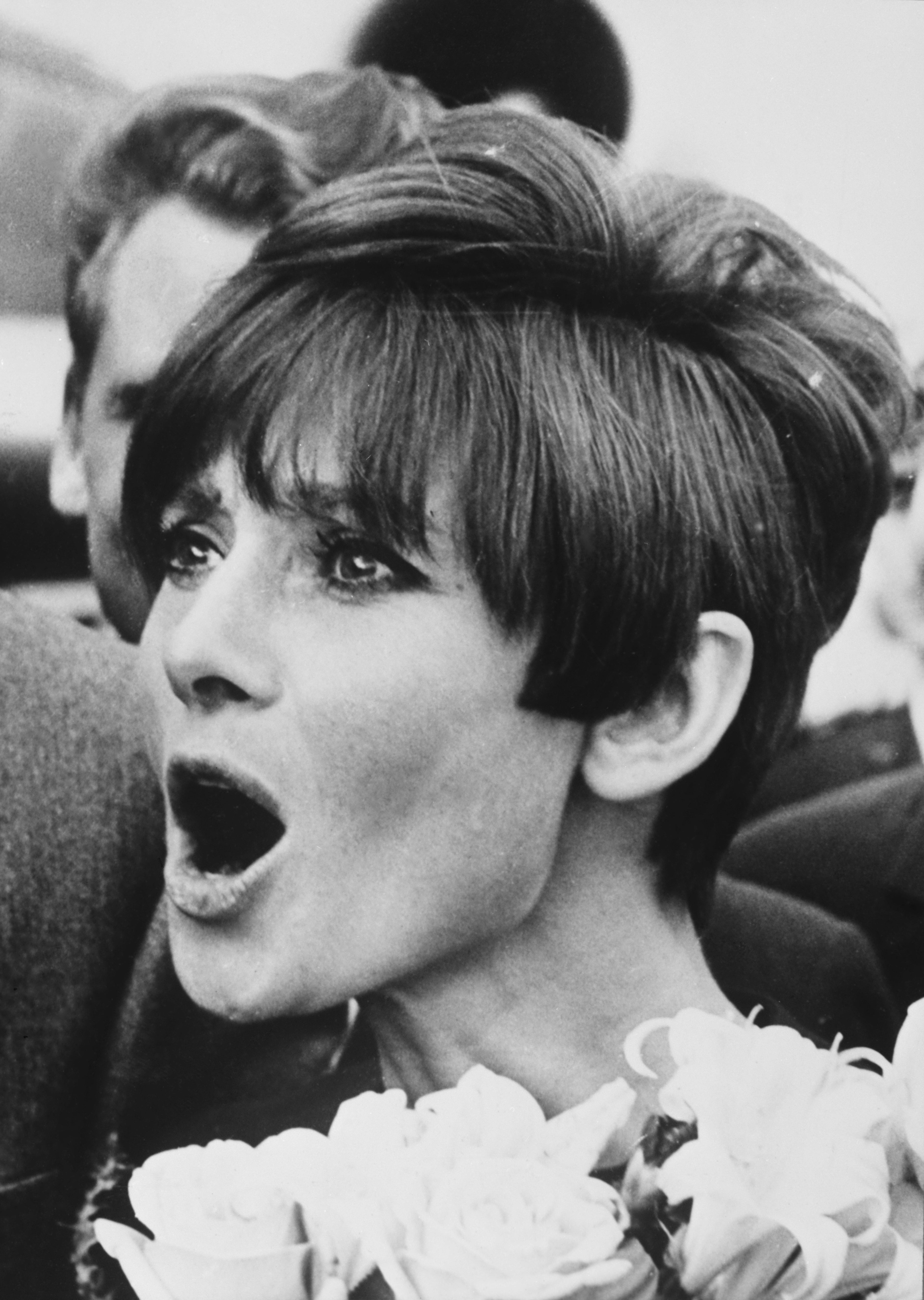Audrey Hepburn with short, stylish hair looks surprised, surrounded by flowers, mouth open in an expressive gesture