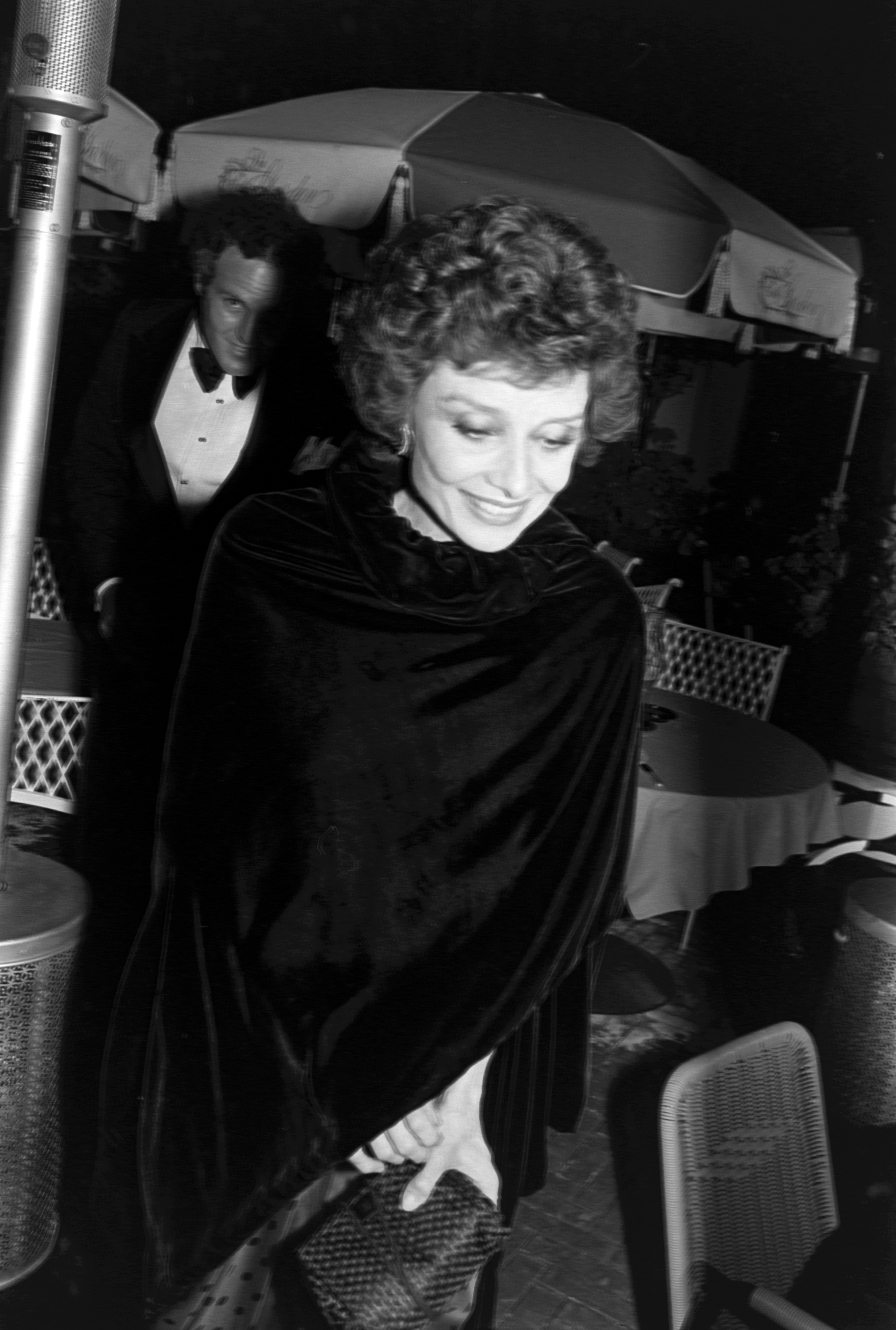 Audrey Hepburn in a formal black velvet cape and dress smiles, walking outdoors at a formal event
