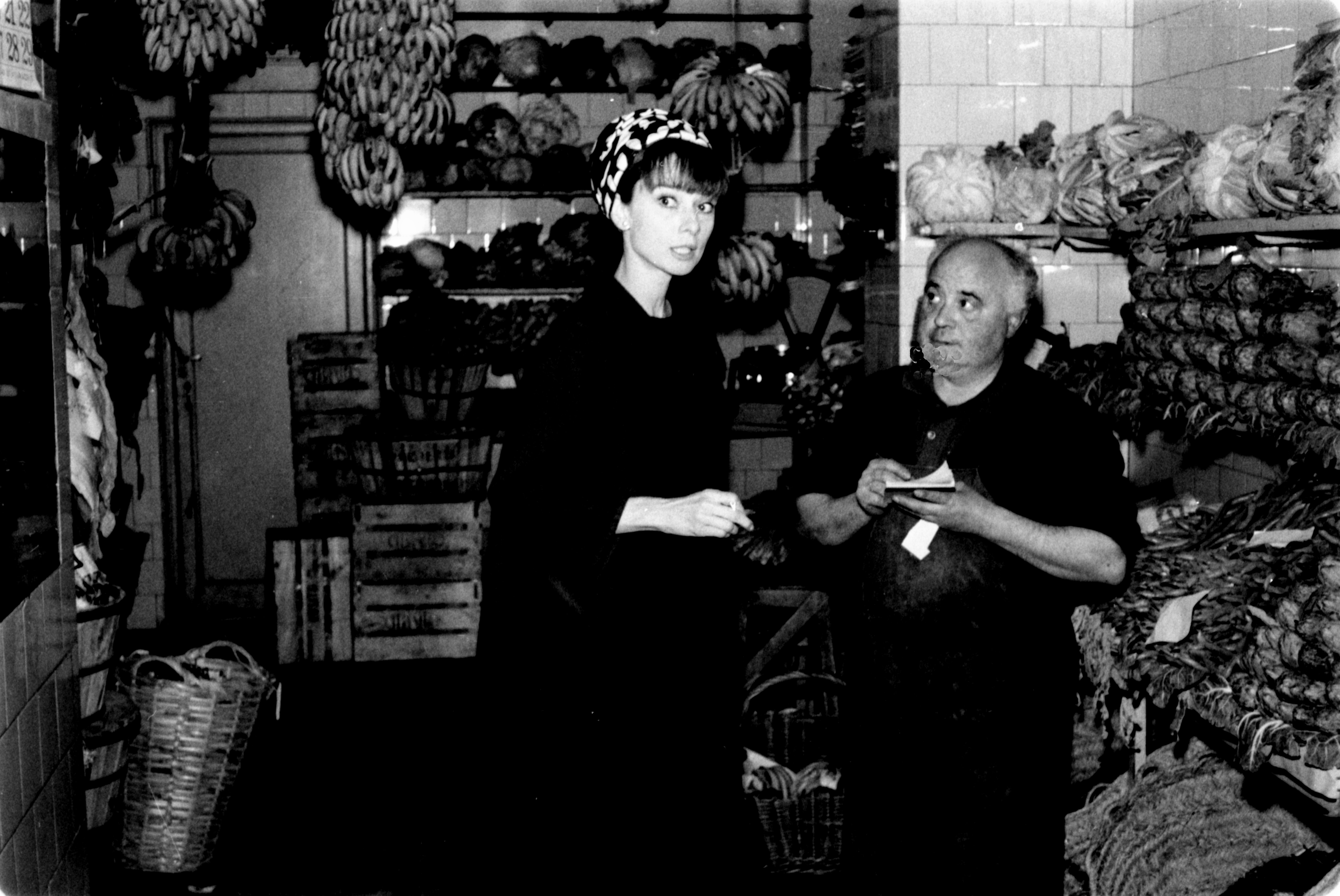 Audrey Hepburn in a chic dress with a headscarf, standing in a market, talking to a vendor surrounded by fruits and vegetables