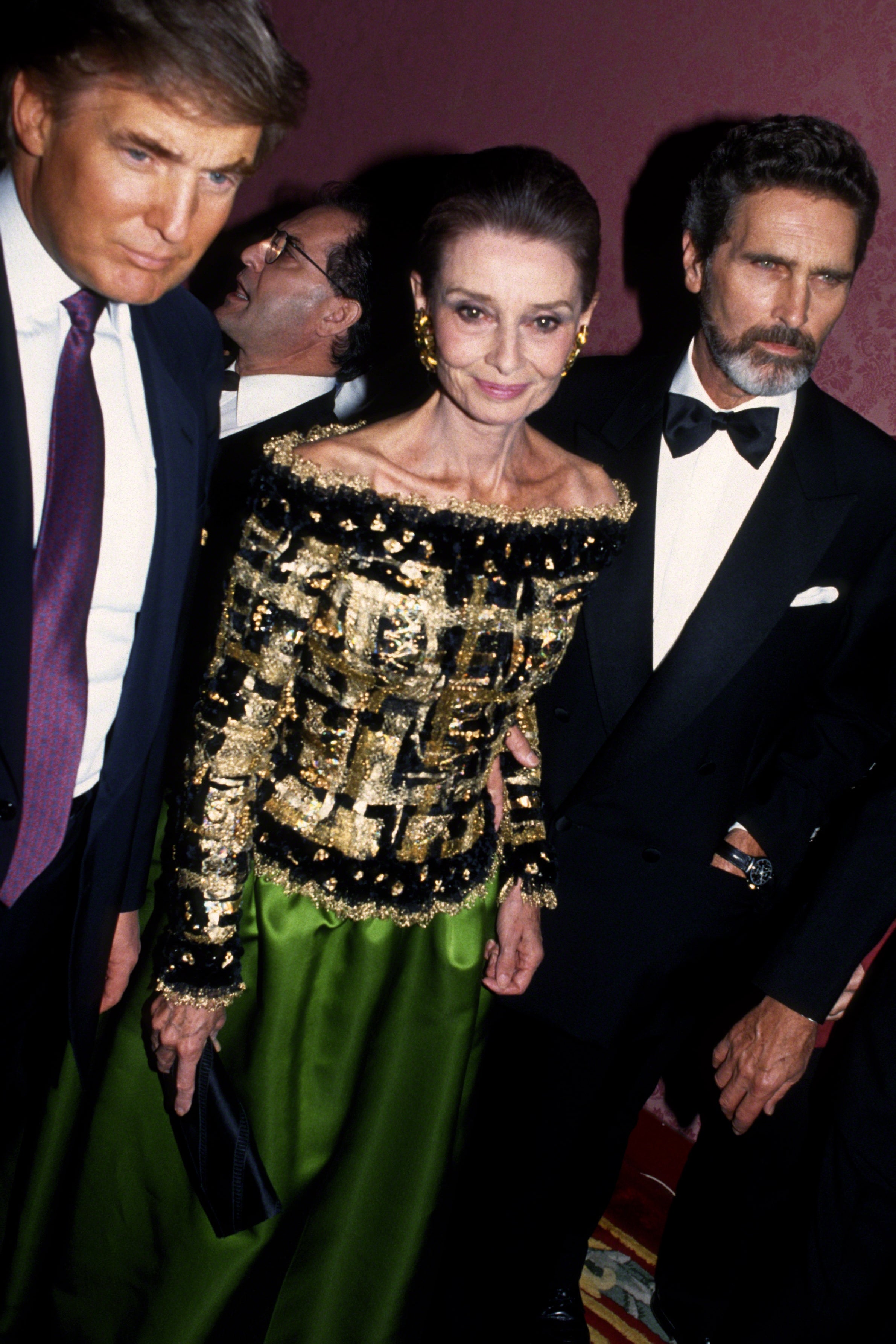 Three people at an event, with one wearing an elegant dress with an ornate top, accompanied by individuals in formal suits