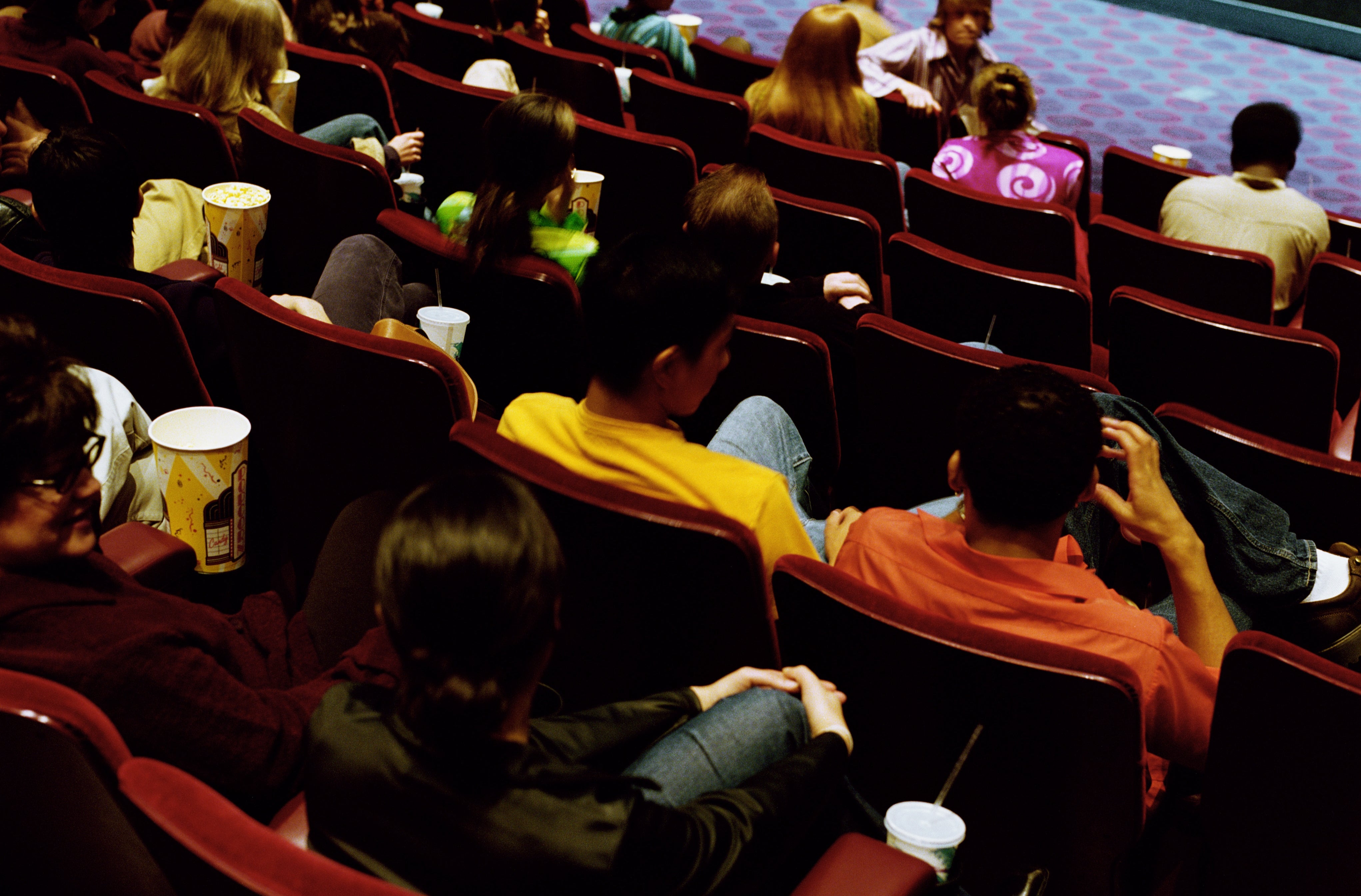 Audience seated in a movie theater, watching a film. People are relaxed with snacks and drinks, focused on the screen ahead