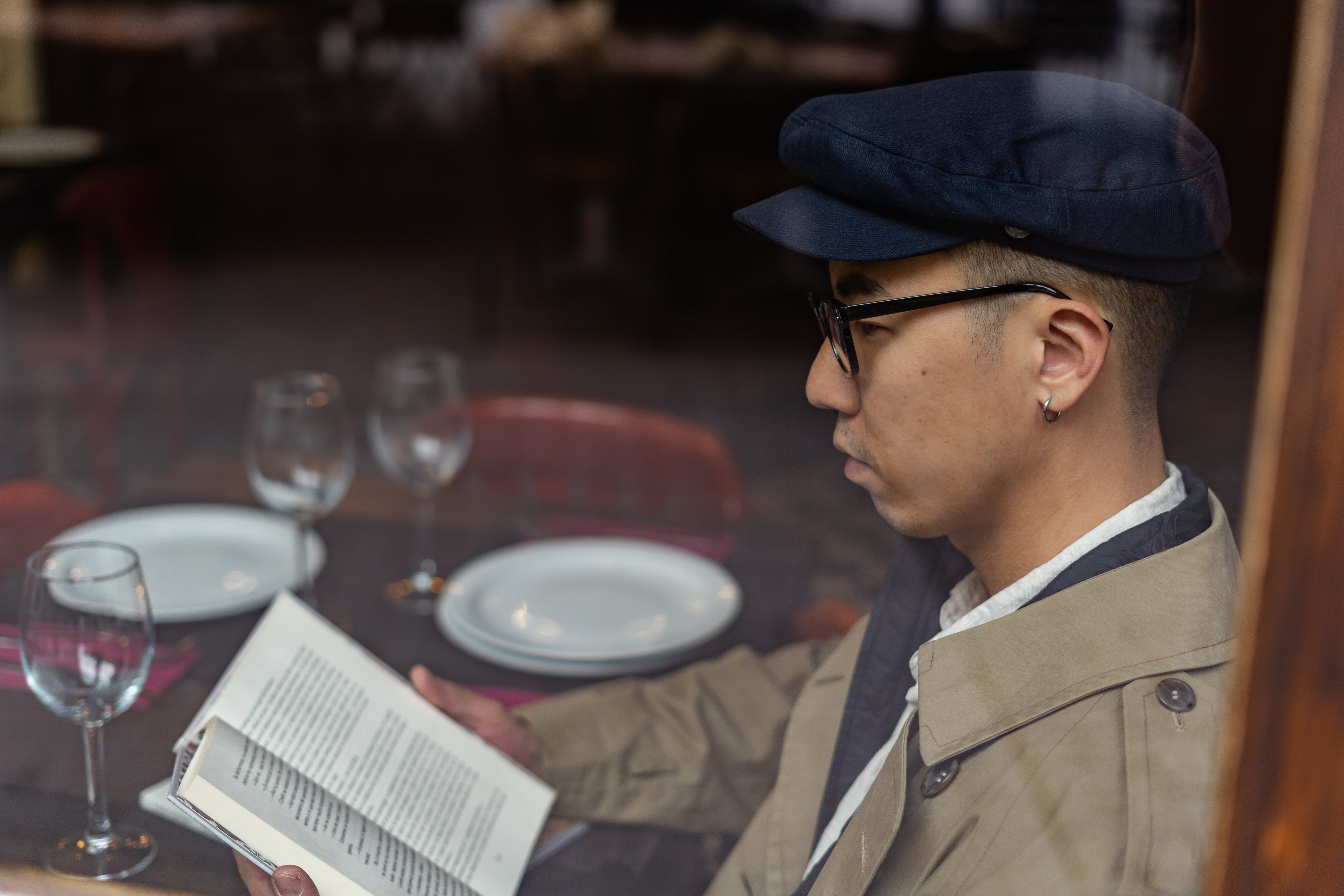 Person sits at a table, reading a book through a window. They wear a flat cap, glasses, and a trench coat. Empty plates and wine glasses are on the table